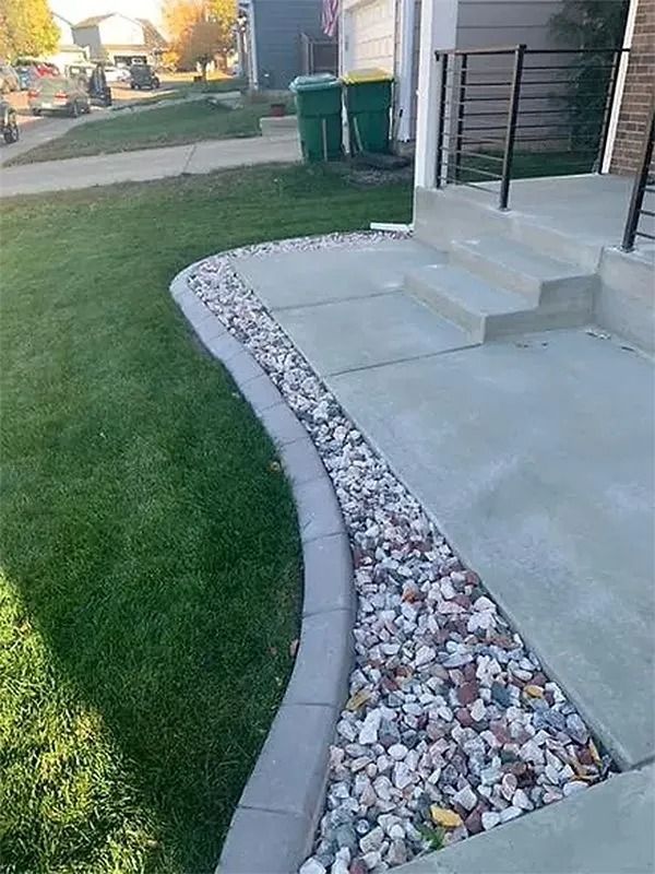 Curved concrete walkway beside a house, bordered by white rocks and green lawn.