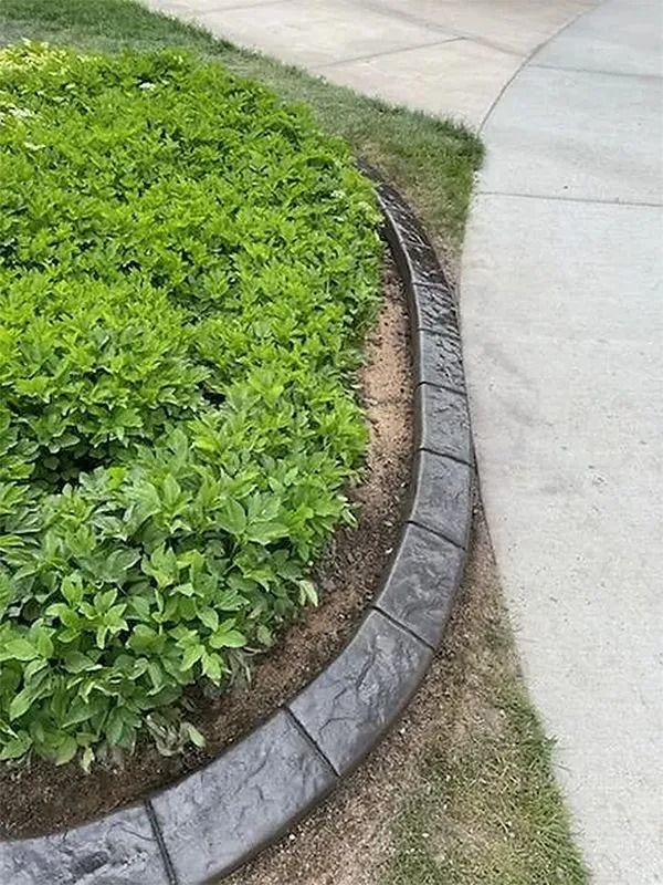 Curved gray edging borders a green shrub bed beside a concrete sidewalk.