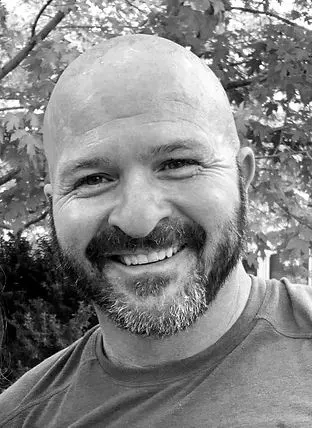 Smiling bald man with a beard outdoors, trees in the background.