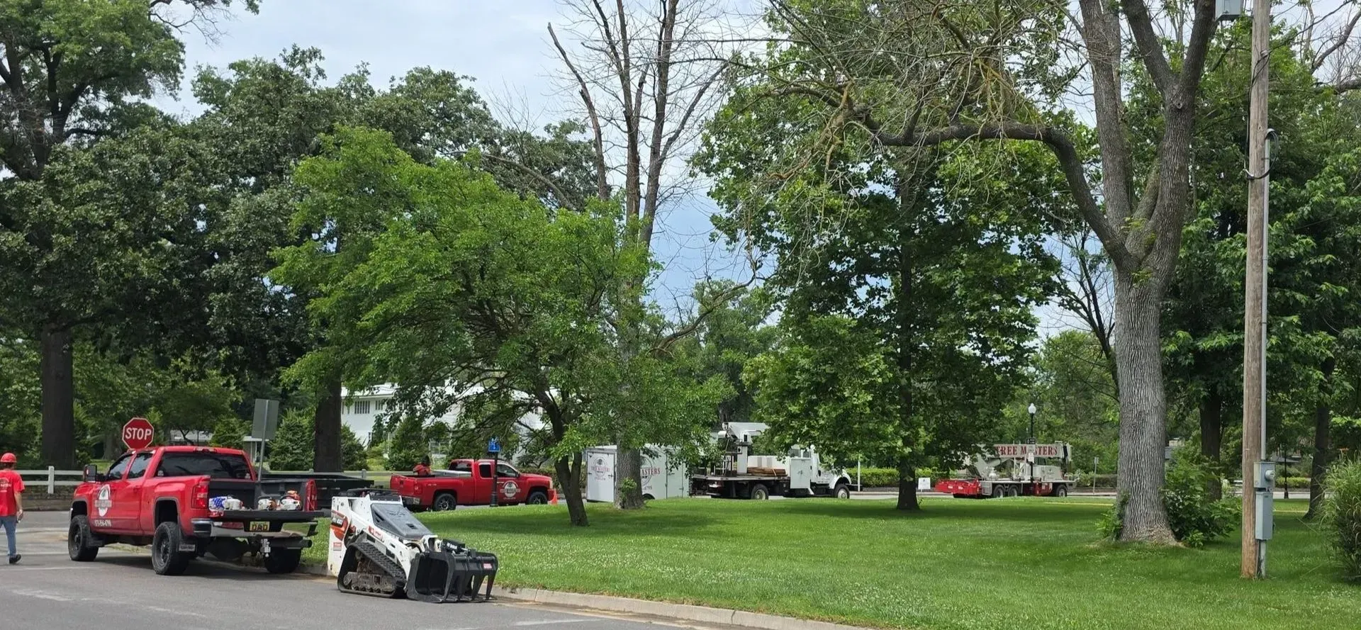 Red fire trucks and a firefighter near a hose in a grassy park with trees and homes in the background