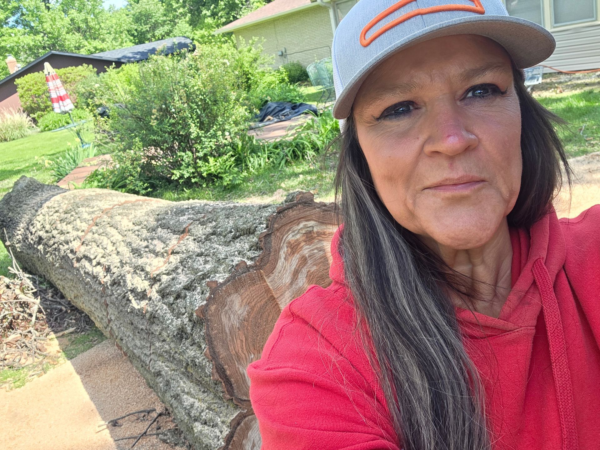 Woman in red hoodie taking a selfie beside a large fallen tree trunk in a leafy yard