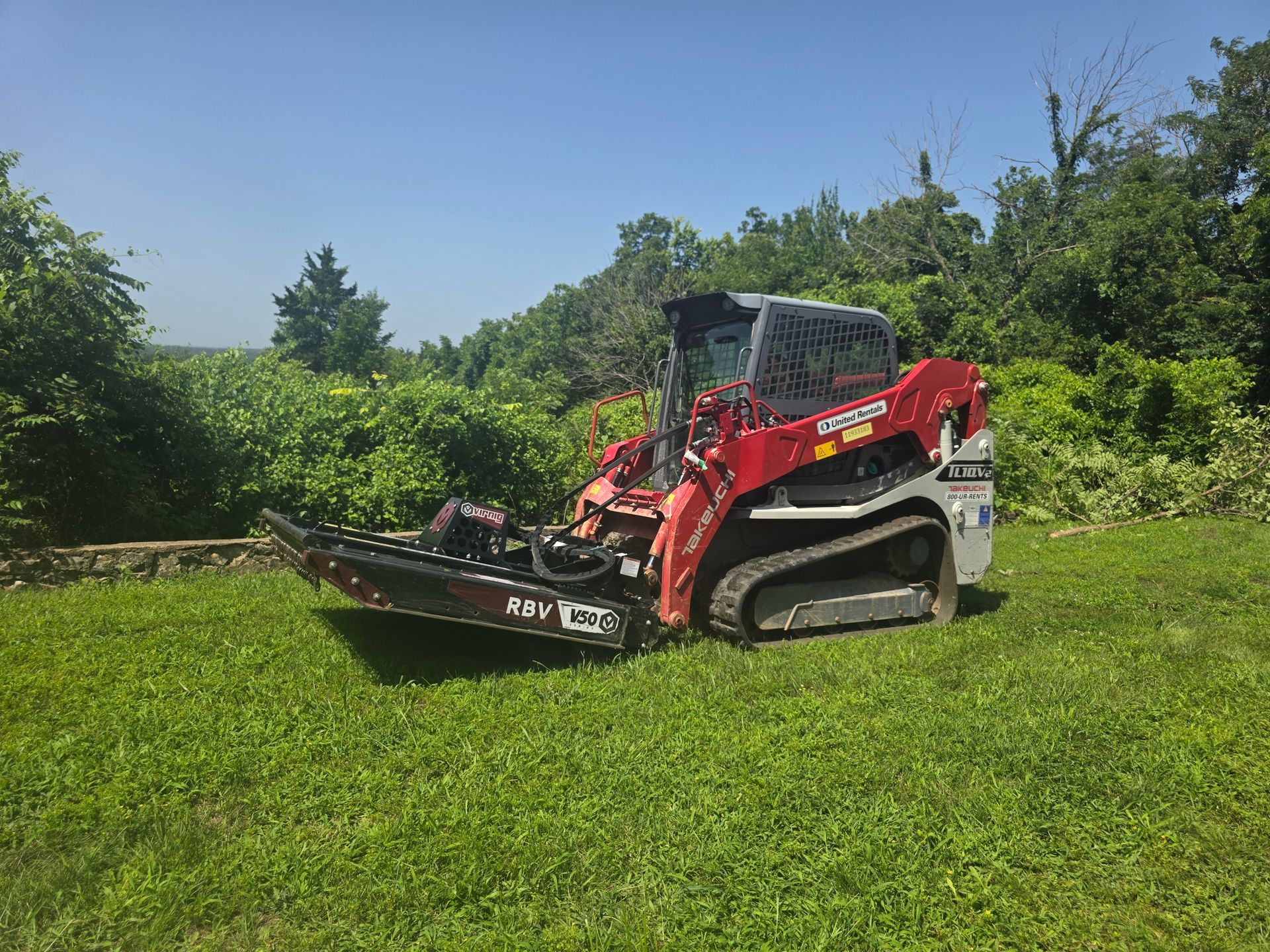 Red skid steer mower on a grassy field beside dense green bushes under a blue sky