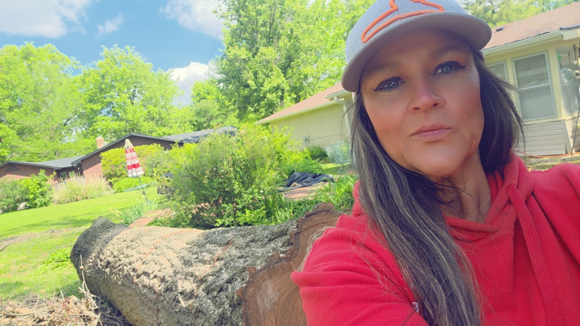 Woman in a red hoodie and cap taking a selfie in a sunny garden with rocks and greenery.