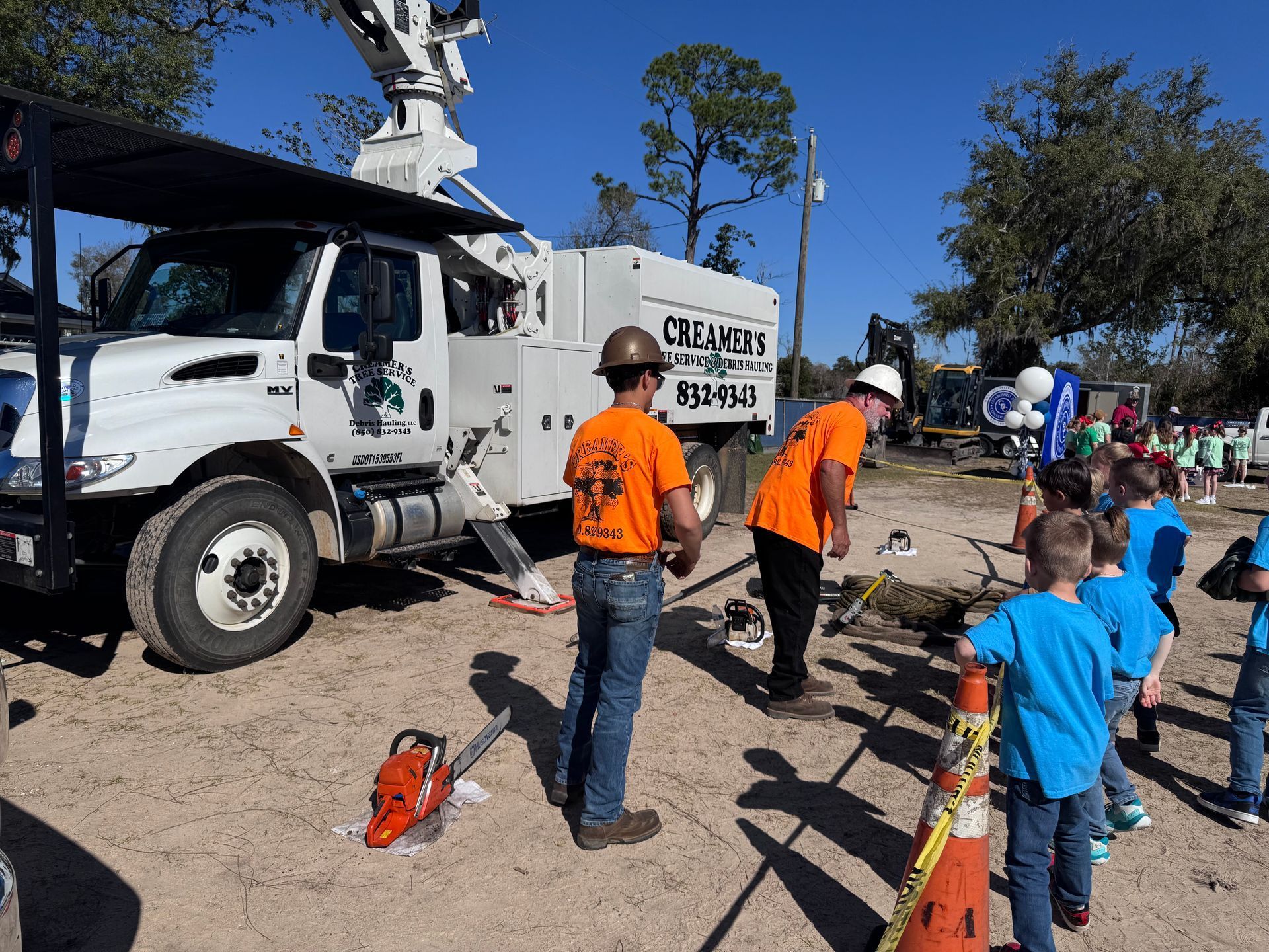 Workers in orange shirts near a white utility truck at an outdoor cleanup site, with children watching nearby