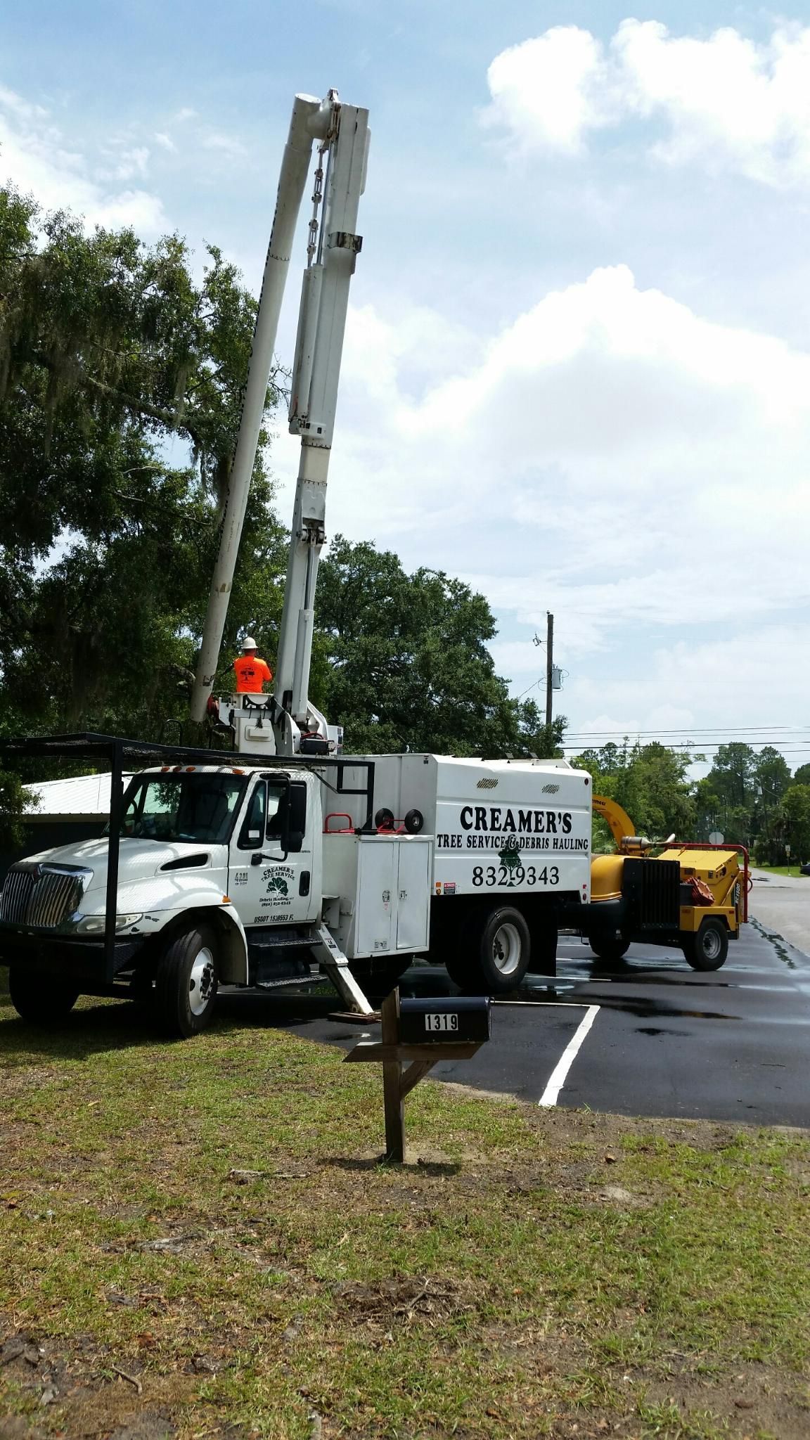 Utility truck with raised boom and worker gear in a parking lot near trees and a yellow machine