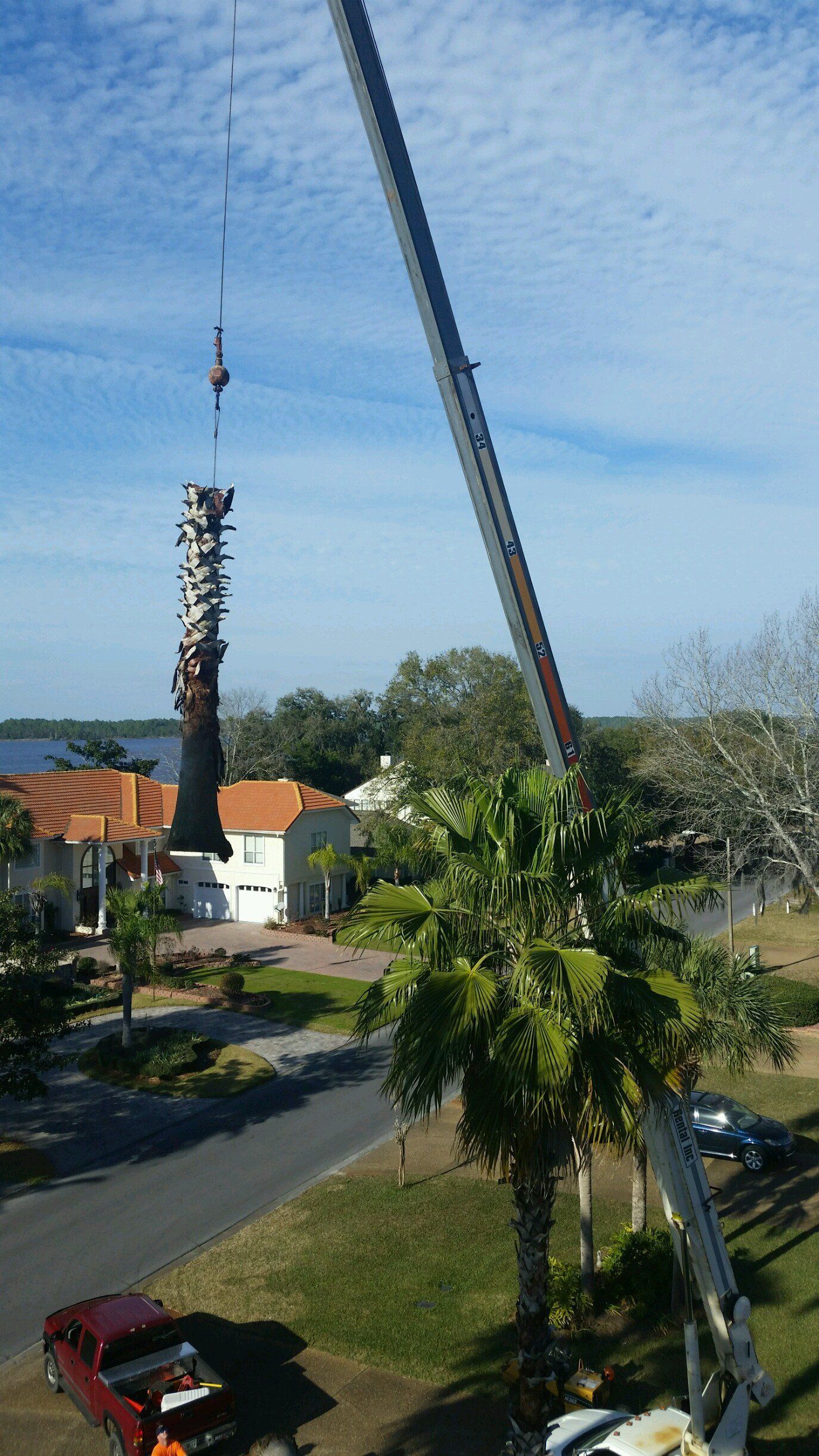 Tall crane lifting a metal tower section beside a road, with houses and trees under a blue sky