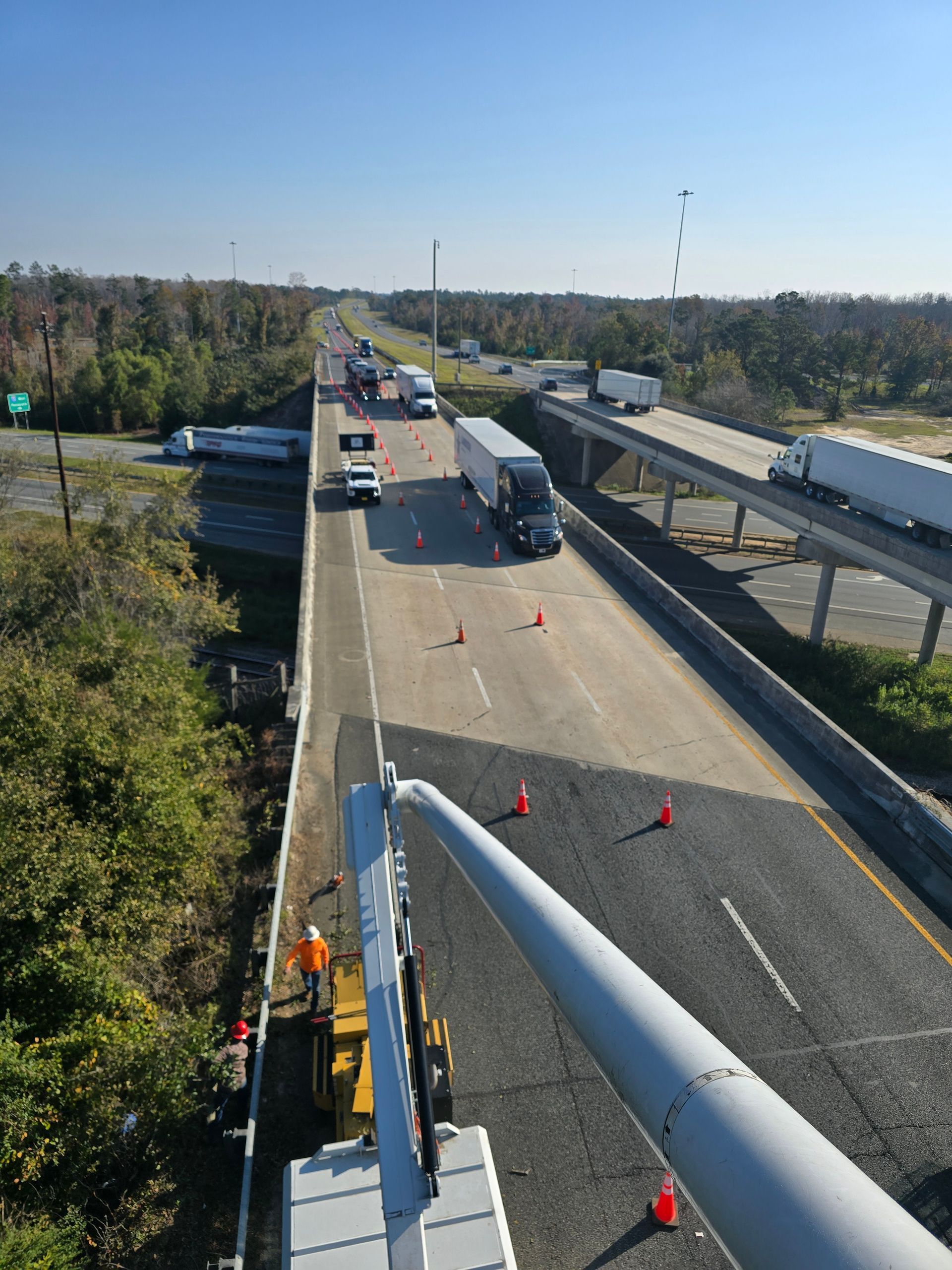 Roadwork on a highway bridge with traffic cones, trucks, and a closed lane under clear sky.