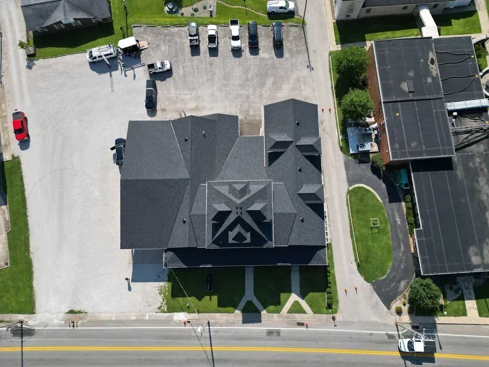 Aerial view of a black-roofed house with a driveway, parked vehicles, and neighboring homes.