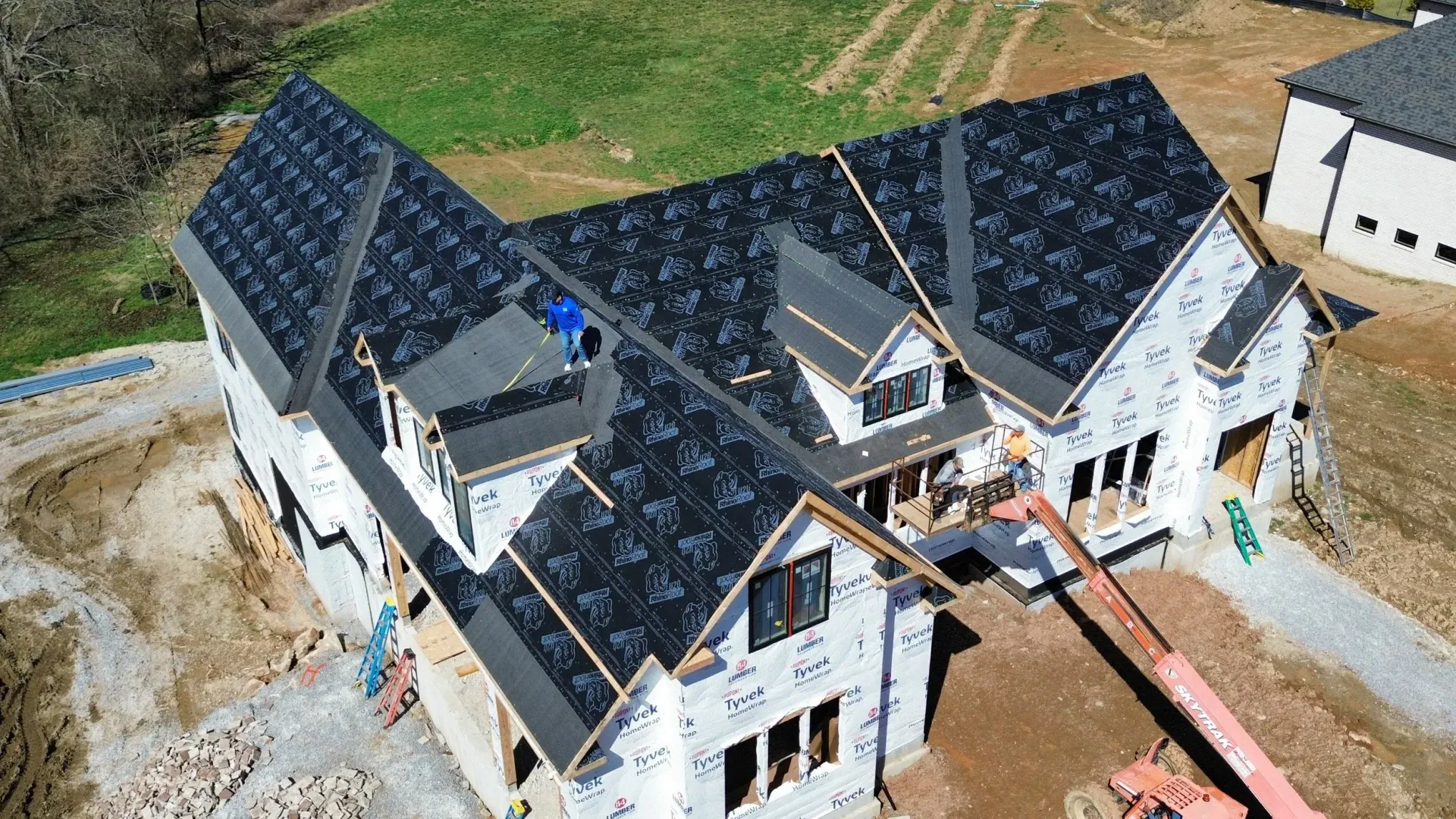 Aerial view of a large house under construction with dark roof shingles and unfinished exterior walls.