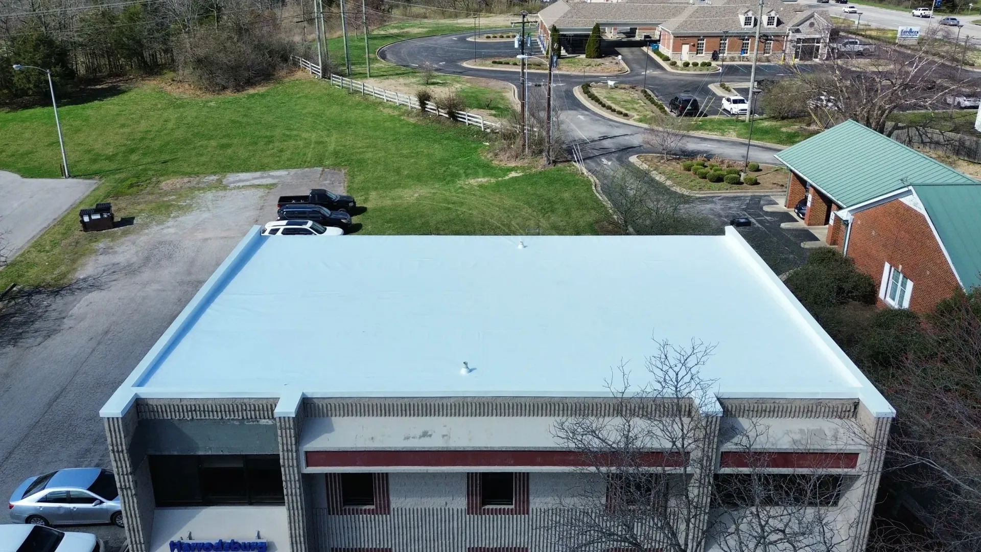 Aerial view of a light-blue roofed building beside a road and grassy area with houses in the background