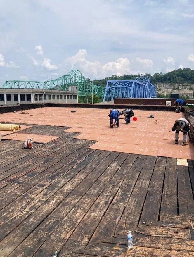 Workers on a rooftop terrace laying tan tiles beside blue steel structures and a green bridge in the distance