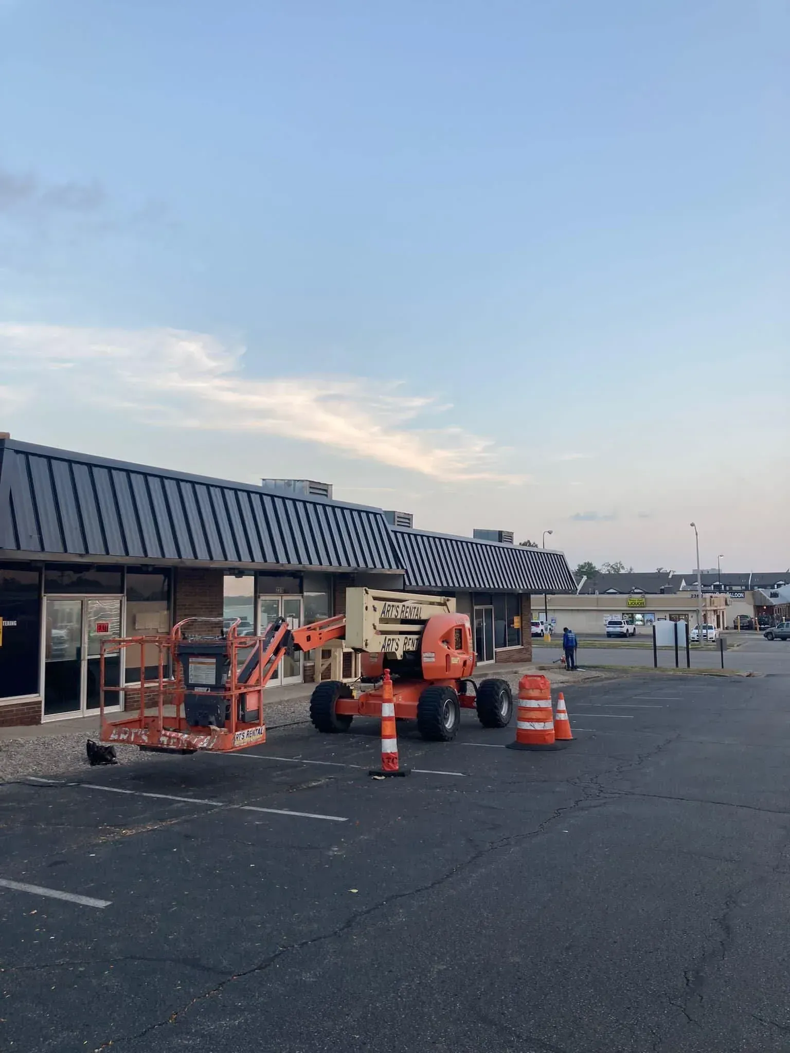 Construction crew working outside a storefront with orange cones, barricades, and equipment in a parking lot at dusk