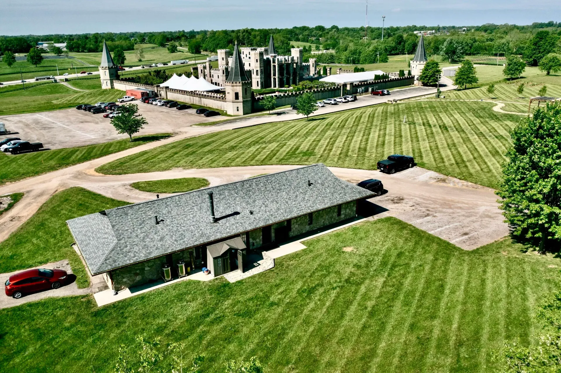 Aerial view of a rural estate with a long building, manicured lawns, trees, and a red car nearby.