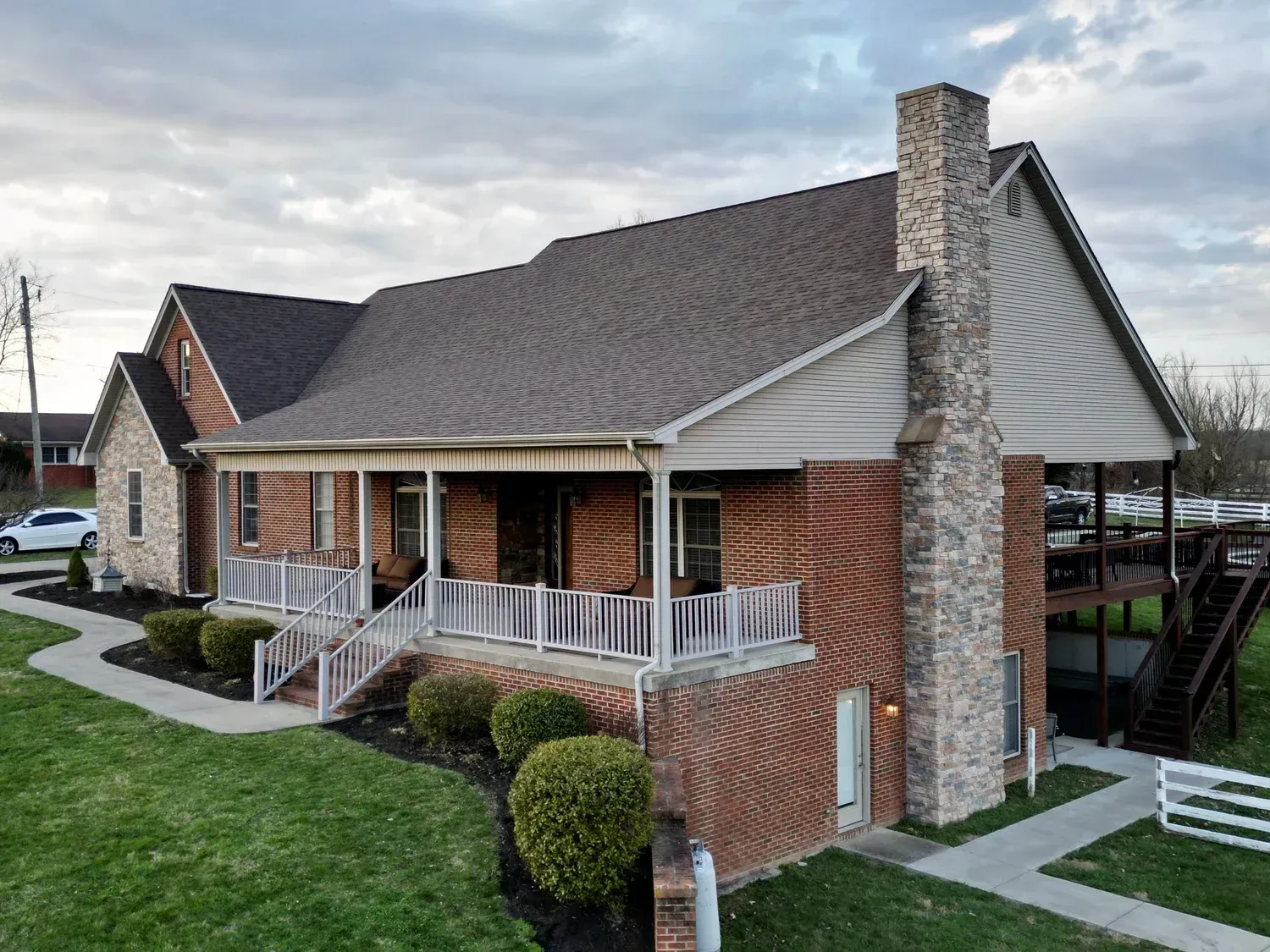 Brick house with gray shingle roof, white porch railings, and stone chimney on a grassy corner lot