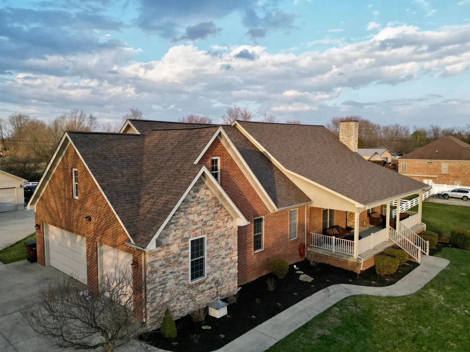 Aerial view of a brick suburban house with a brown roof, front porch, and attached garage under a cloudy sky