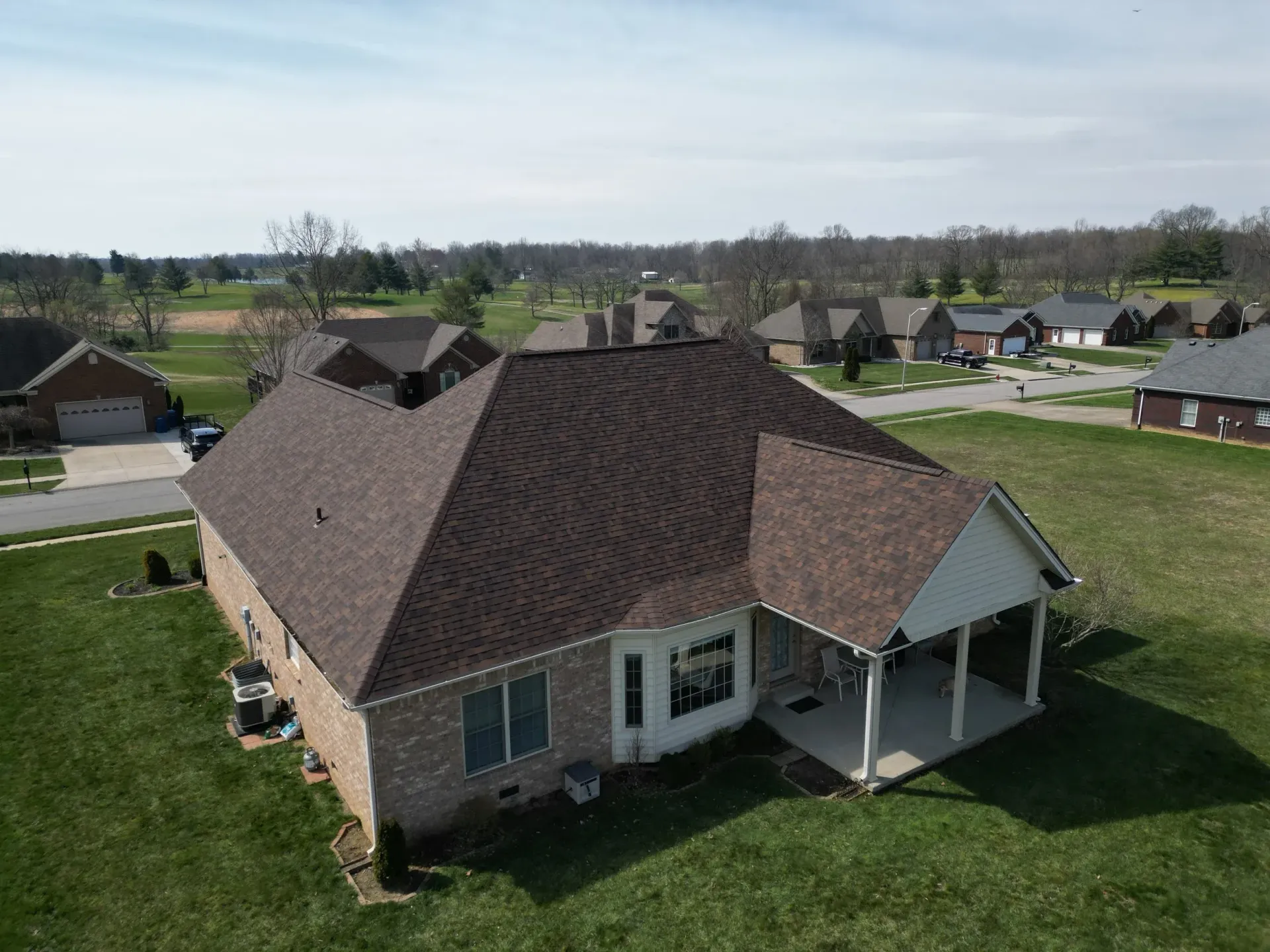 Aerial view of a brick house with a brown roof and covered porch in a suburban neighborhood