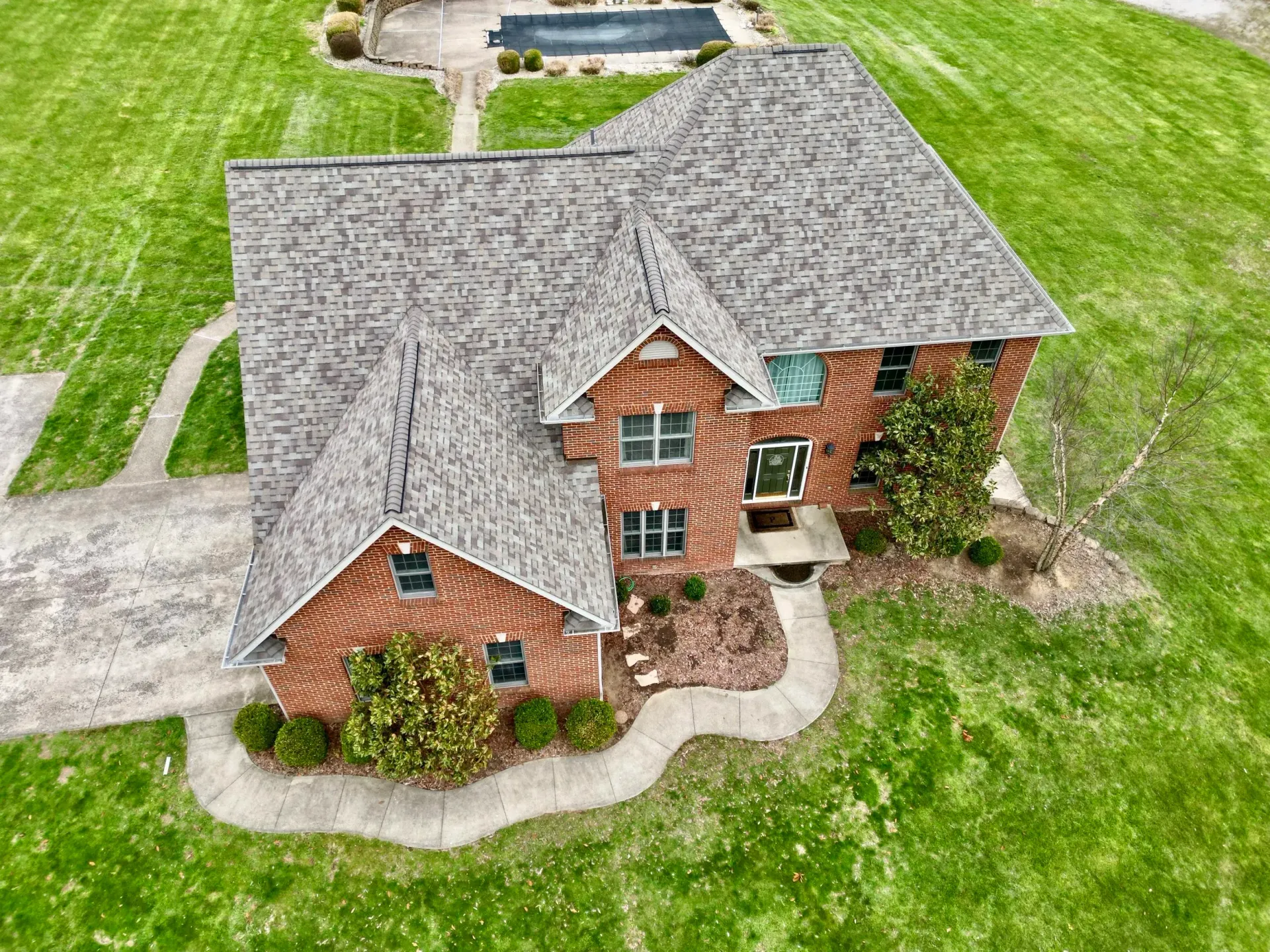 Aerial view of a large brick house with a gray shingled roof and a curved walkway in a grassy yard.