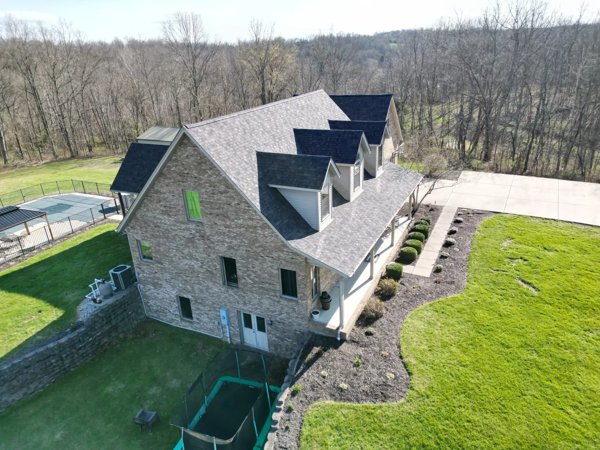 Aerial view of a stone house with dark roof, green lawn, and wooded surroundings
