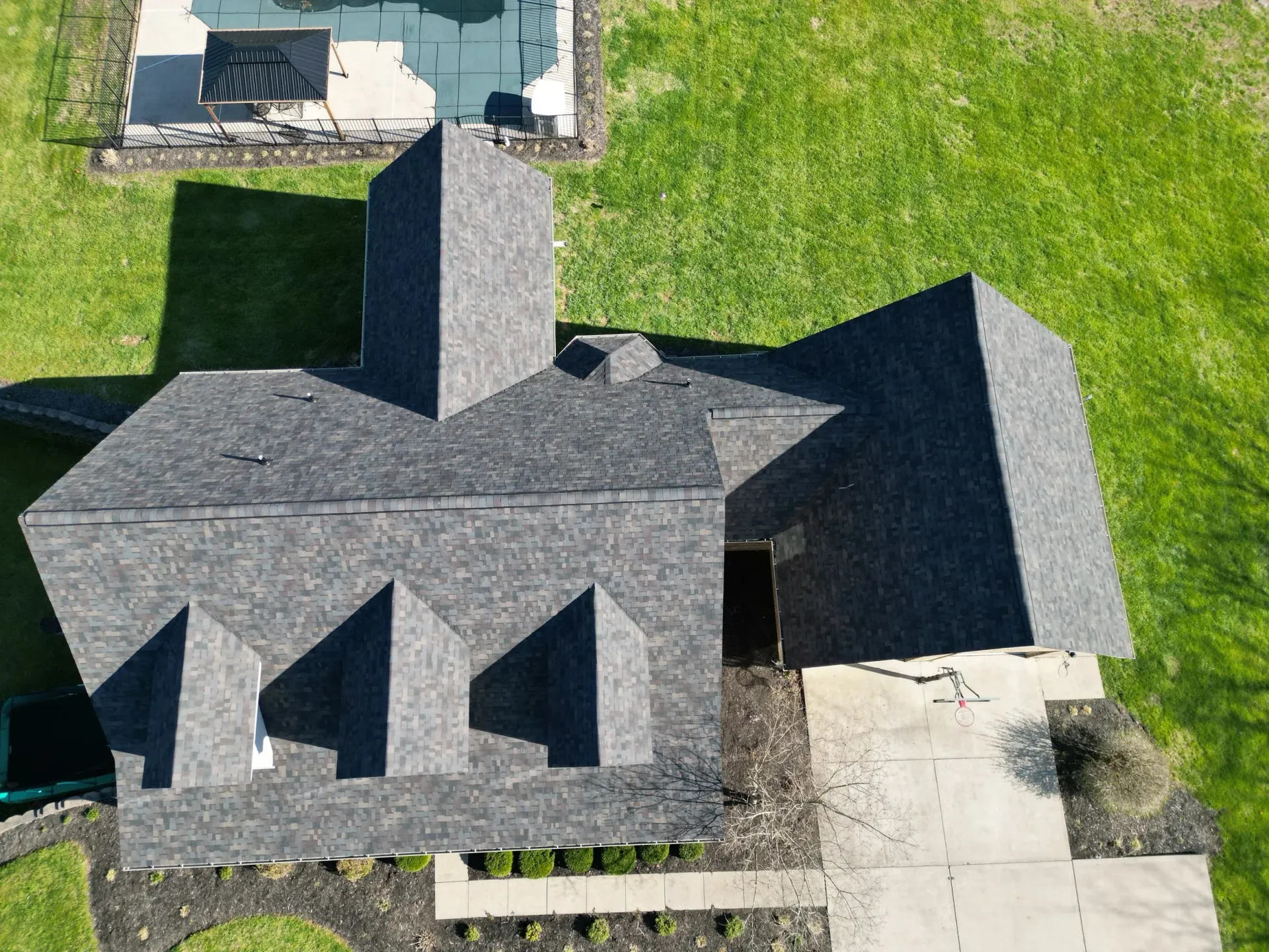 Aerial view of a gray-roofed house with dormer windows, chimney, driveway, and green lawn.
