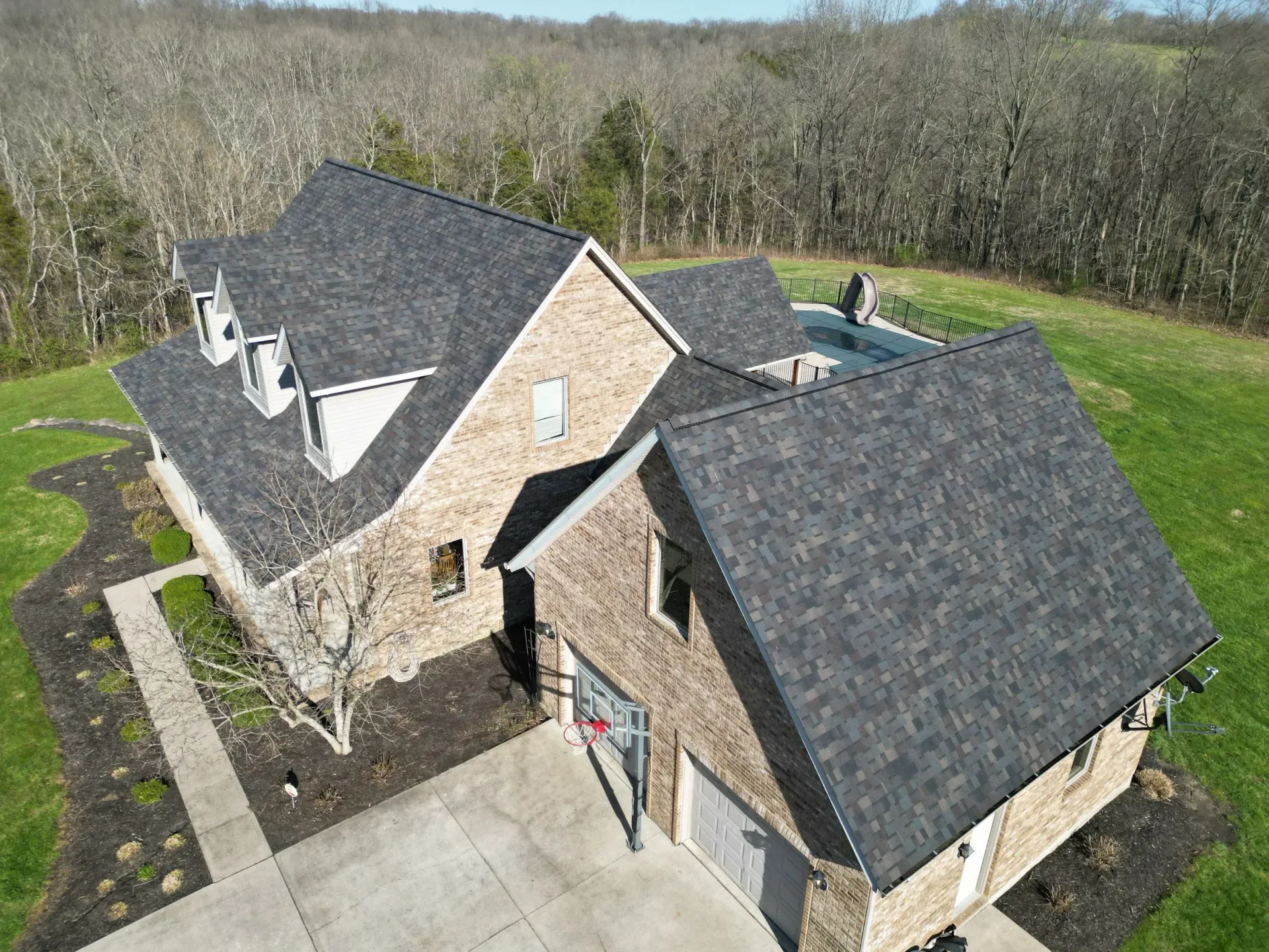 Aerial view of a stone house with dark roof, front driveway, and a backyard pool beside a wooded area
