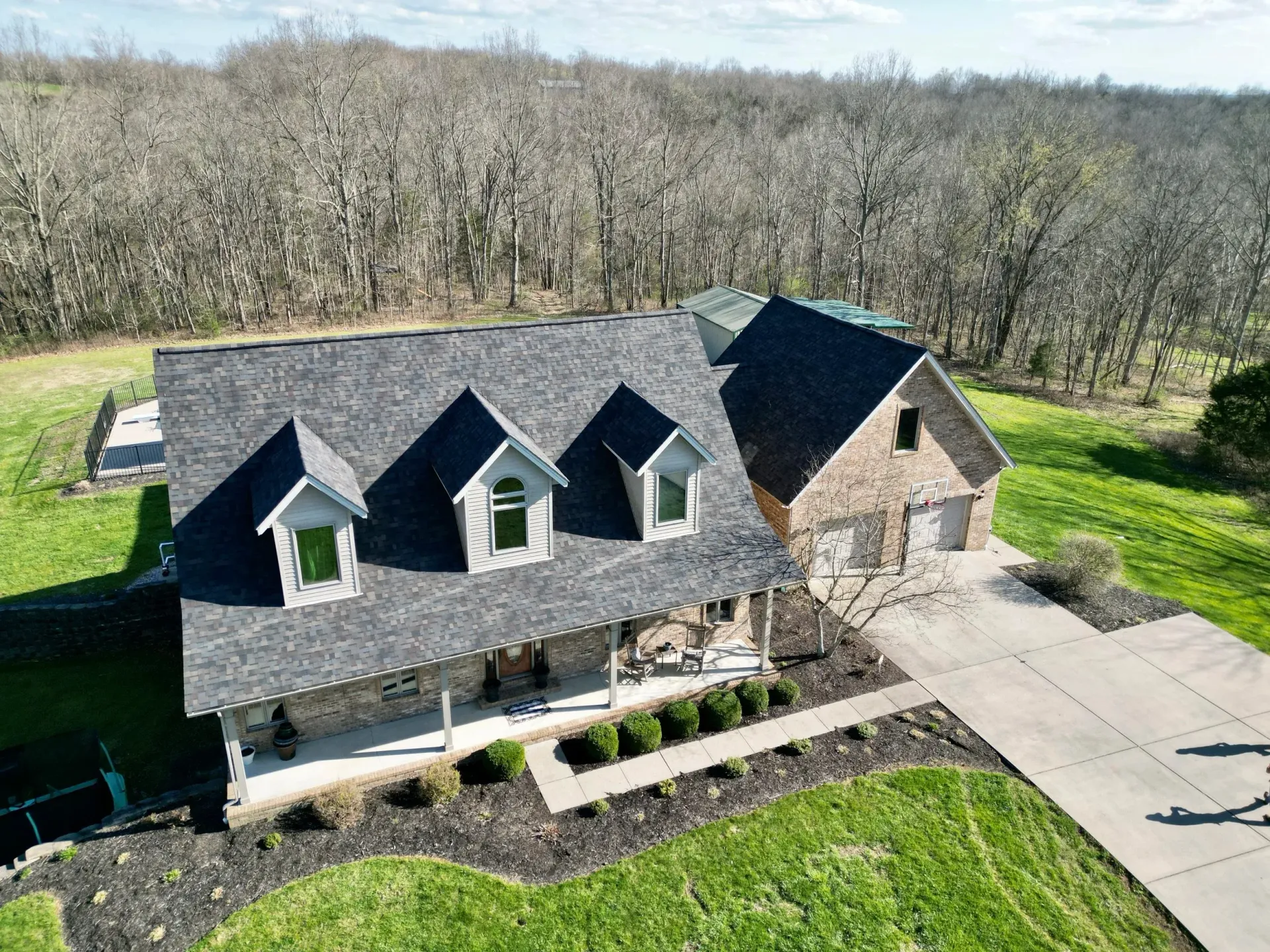 Aerial view of a gray-roofed house with a stone garage, driveway, and wooded surroundings.
