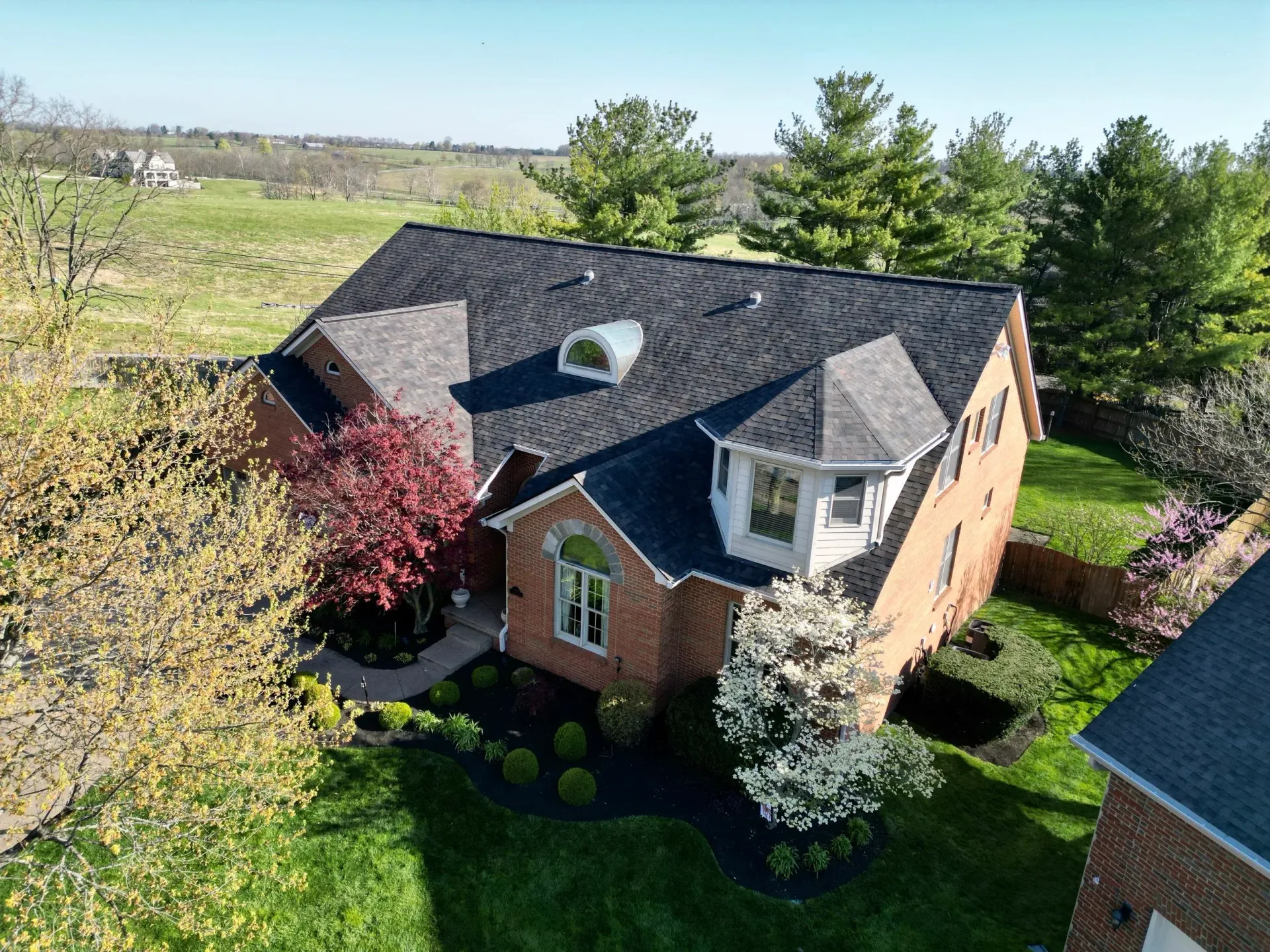 Brick suburban house with black roof, dormer windows, and landscaped yard under a sunny sky