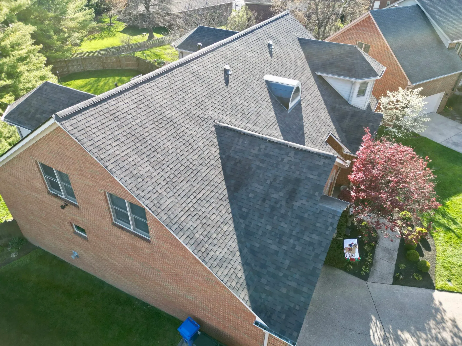 Aerial view of a brick house with a dark shingled roof, driveway, and a pink flowering tree in the yard