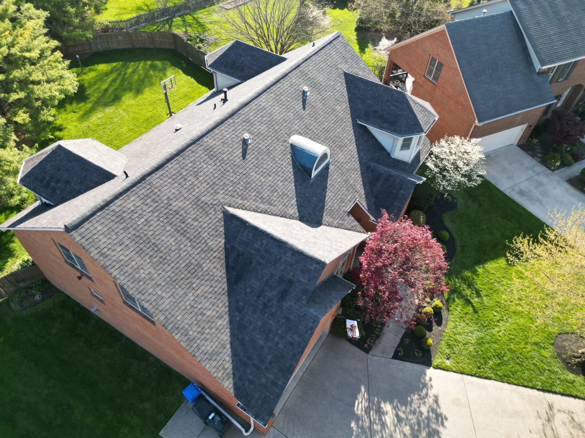 Aerial view of a brick house with a gray shingled roof, skylights, and a pink flowering tree in the yard