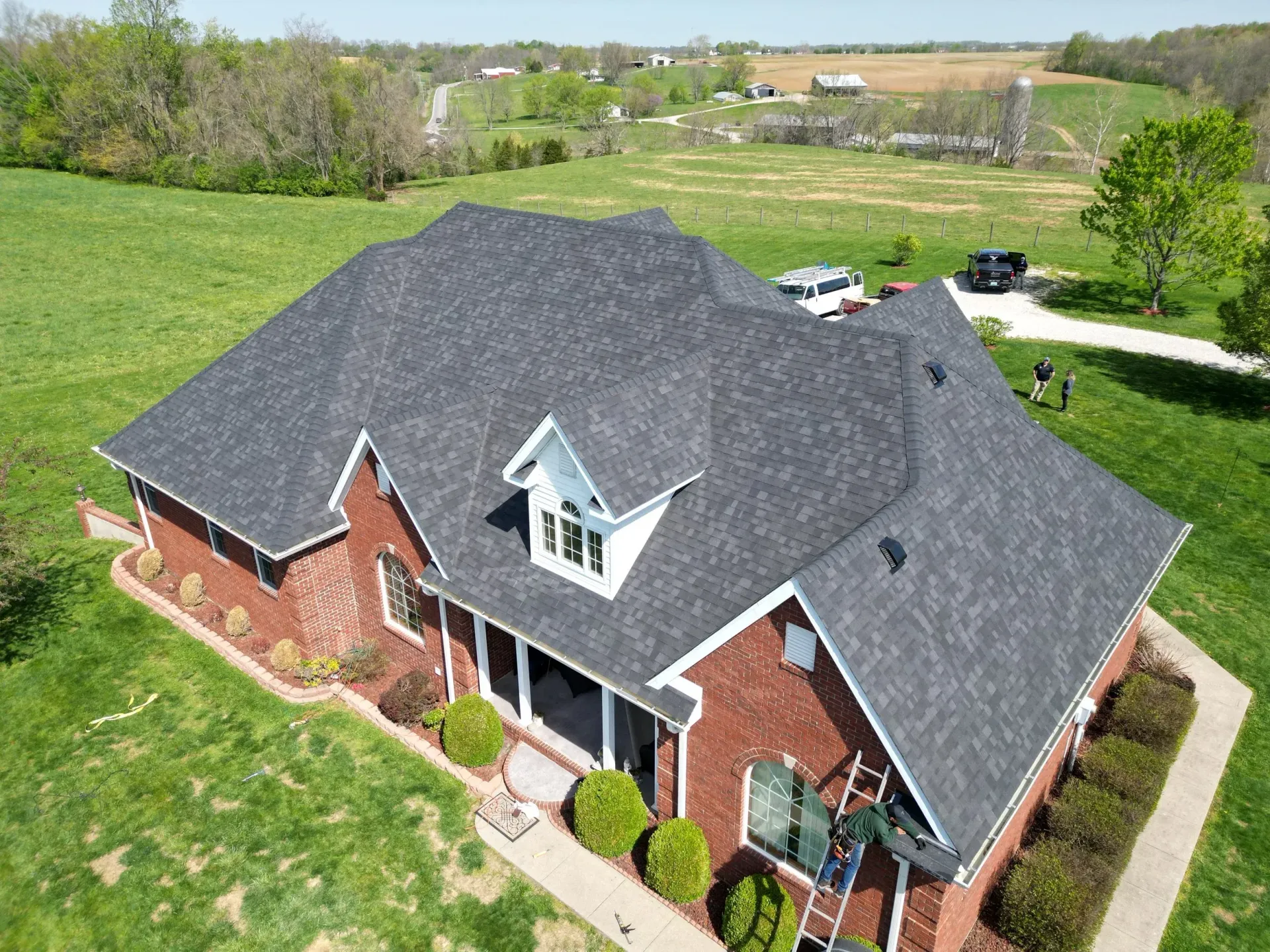 Aerial view of a red brick house with a dark gray roof, surrounded by a green lawn and countryside.