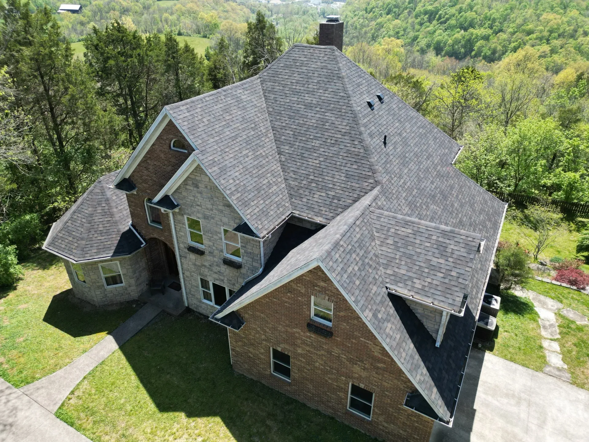 Aerial view of a large brick house with a gray roof, surrounded by green trees and lawn