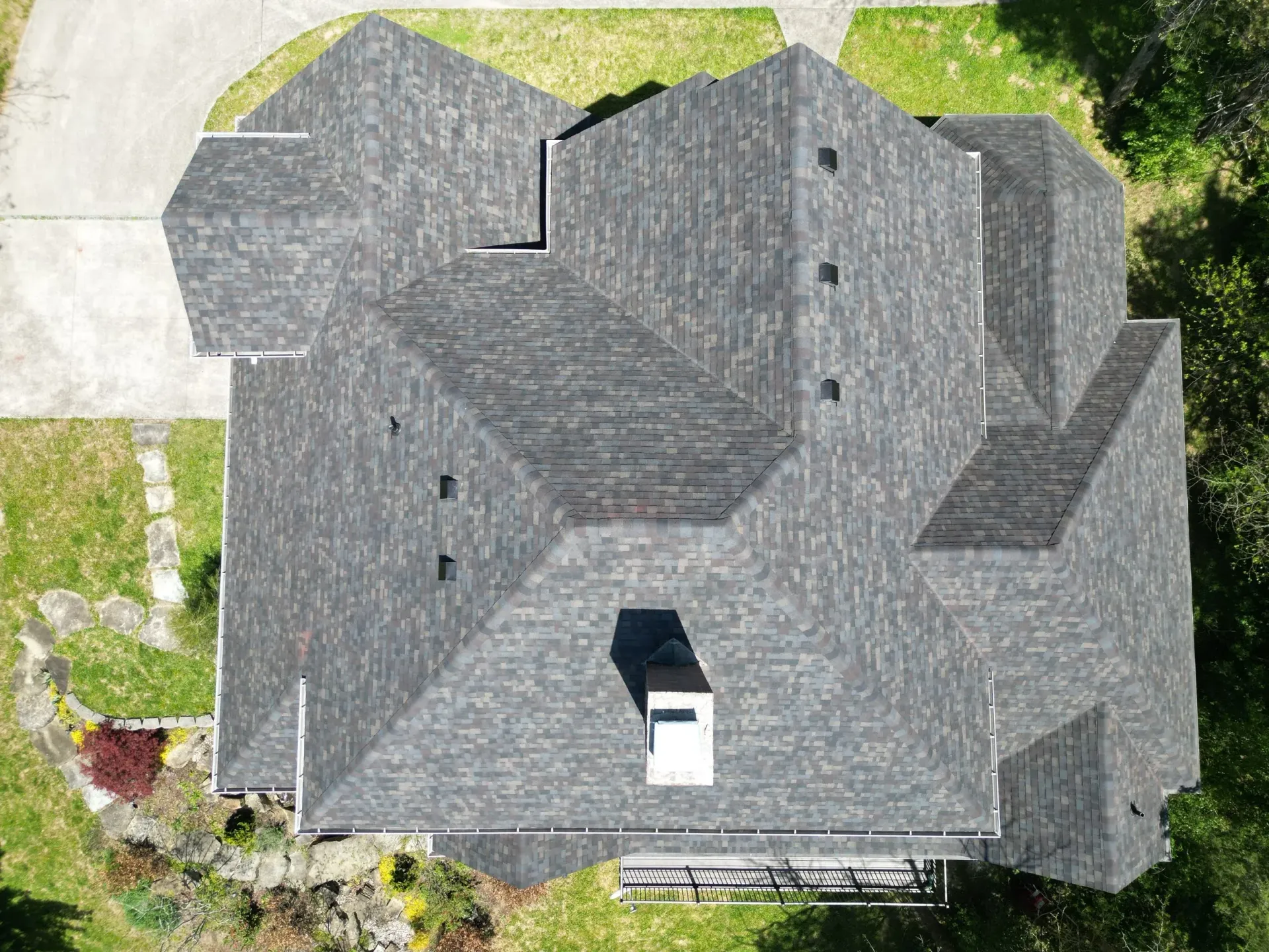 Aerial view of a gray-roofed house with a white chimney surrounded by grass and a driveway.