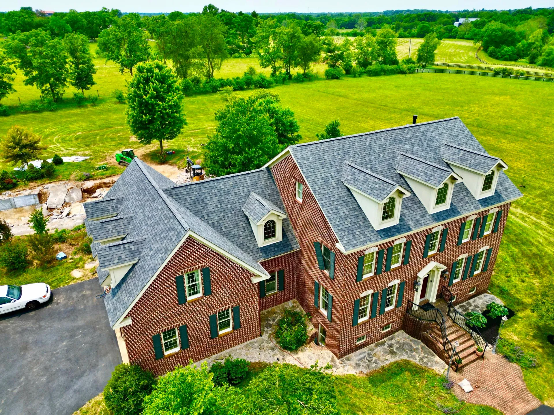 Red brick house with gray roof beside a driveway, surrounded by green fields and trees.