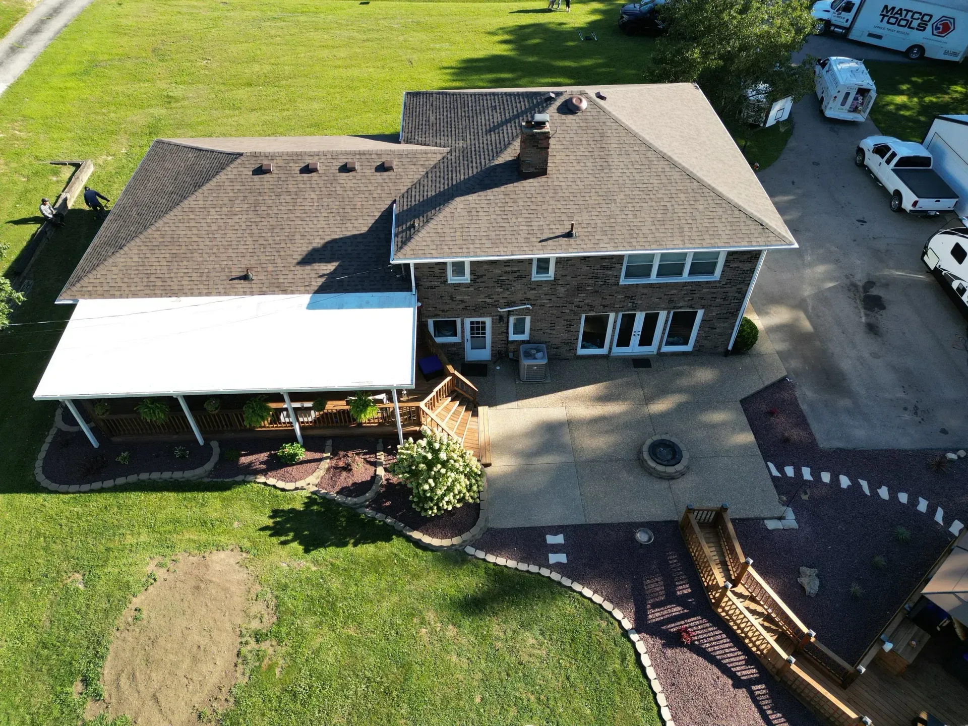 Aerial view of a brick house with a shingled roof, white patio cover, and a landscaped backyard.