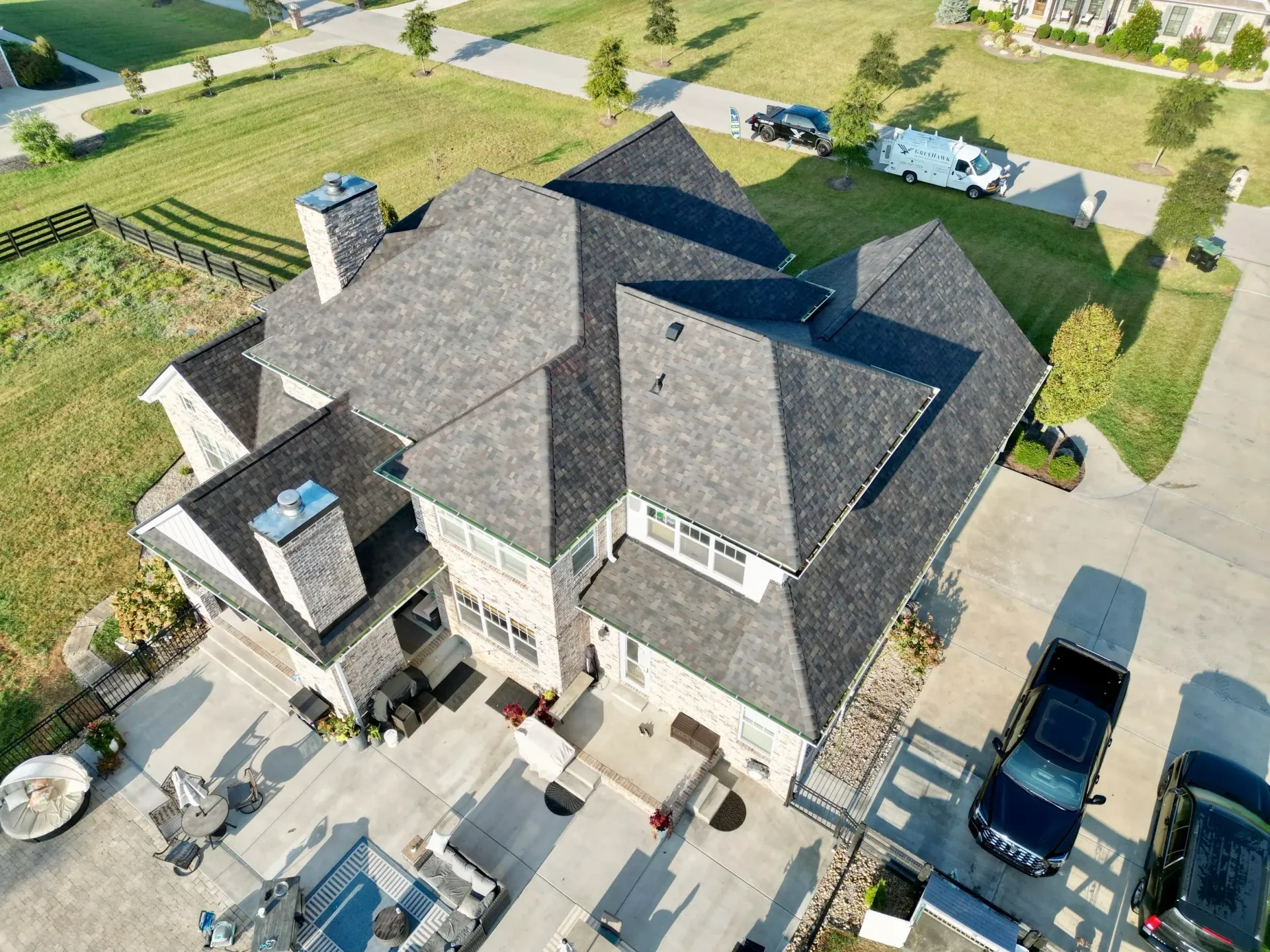 Aerial view of a large gray-roofed house with a driveway, parked vehicles, and surrounding lawns.