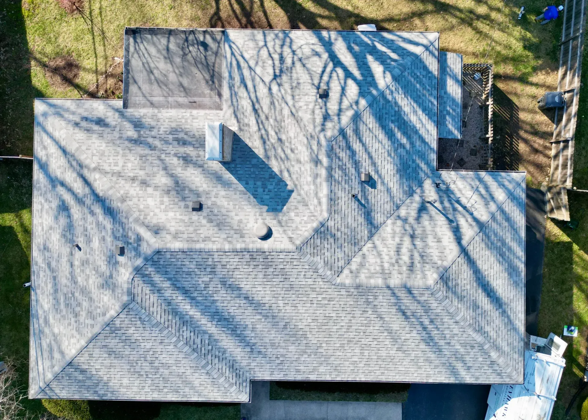 Aerial view of a gray shingled house roof with tree shadows and a small vent on top.