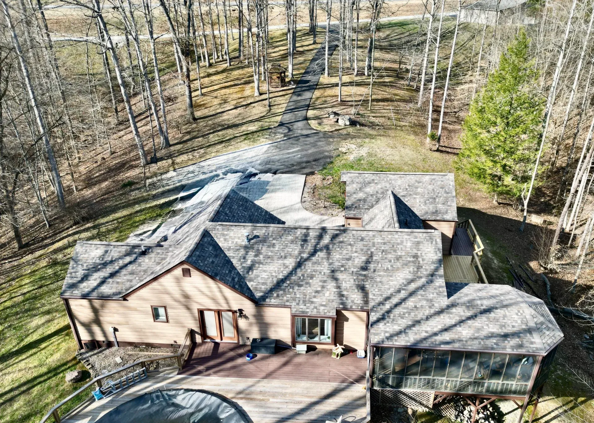 Aerial view of a house with gray shingles in a wooded area, with a driveway and parked vehicle.