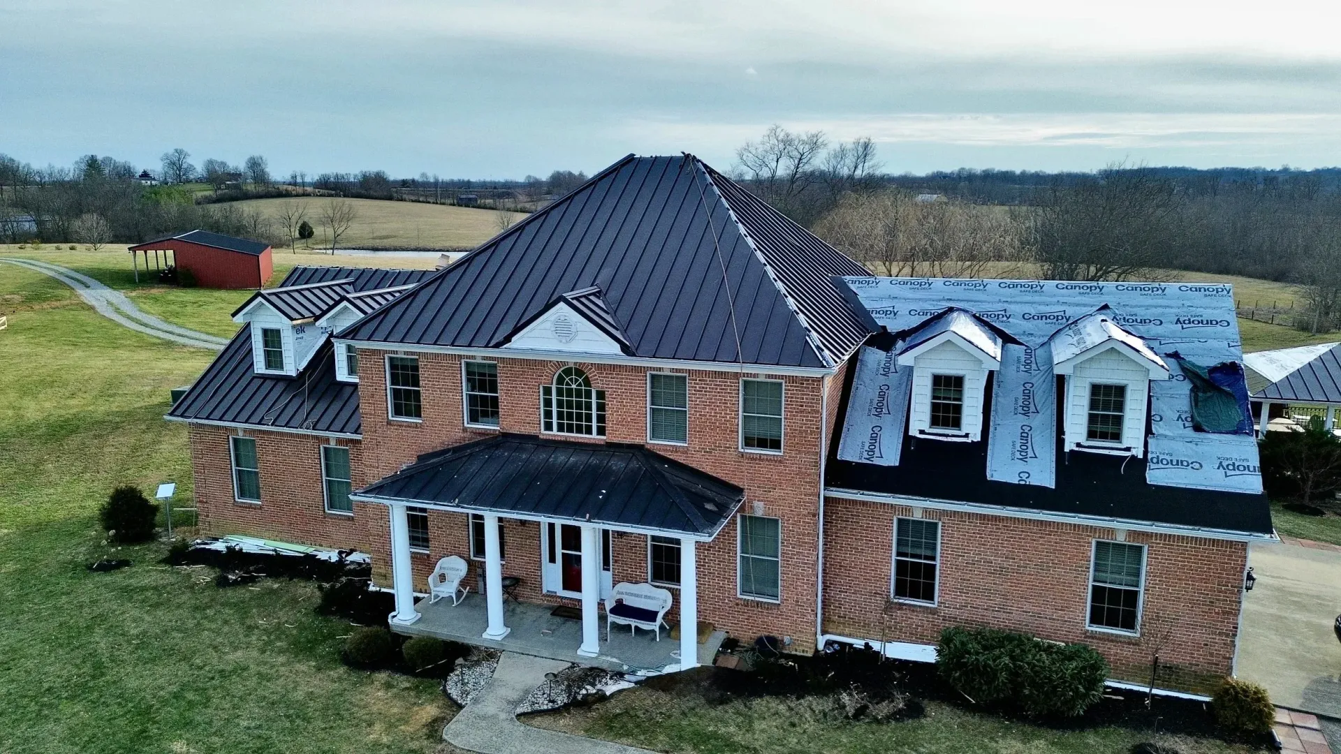Large brick suburban house with a dark roof, front porch, and dormer windows on a grassy lot