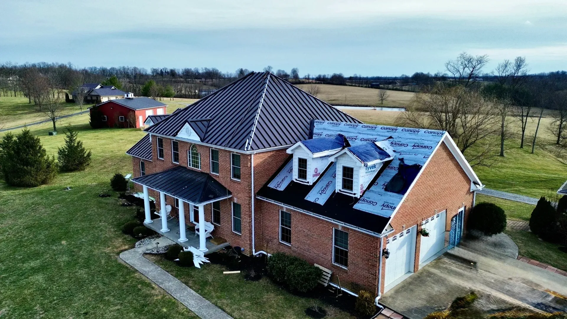 A brick house with a dark roof and white porch sits in a grassy rural field.