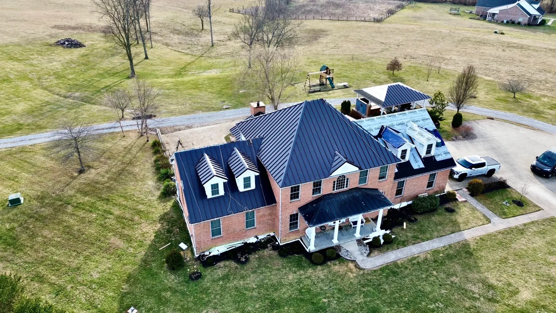 Aerial view of a brick house with a dark roof, white porch, and surrounding grassy yard on a rural road