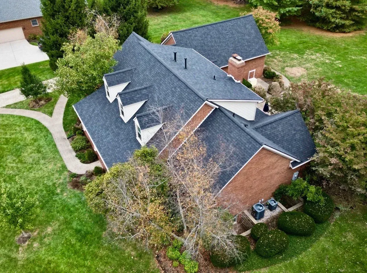 Aerial view of a large house with a dark roof, brick walls, and a green yard surrounded by trees.