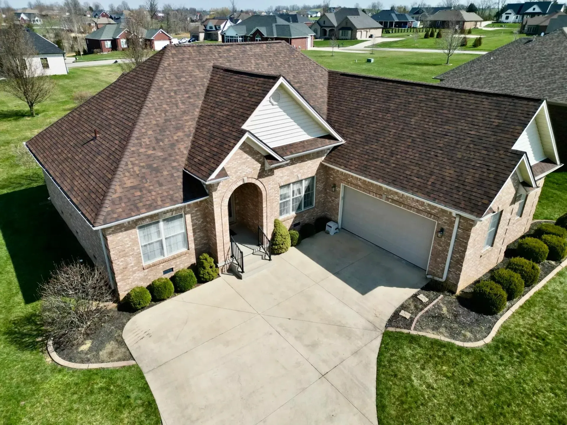 Aerial view of a beige brick suburban house with a brown roof, two-car garage, and curved driveway.