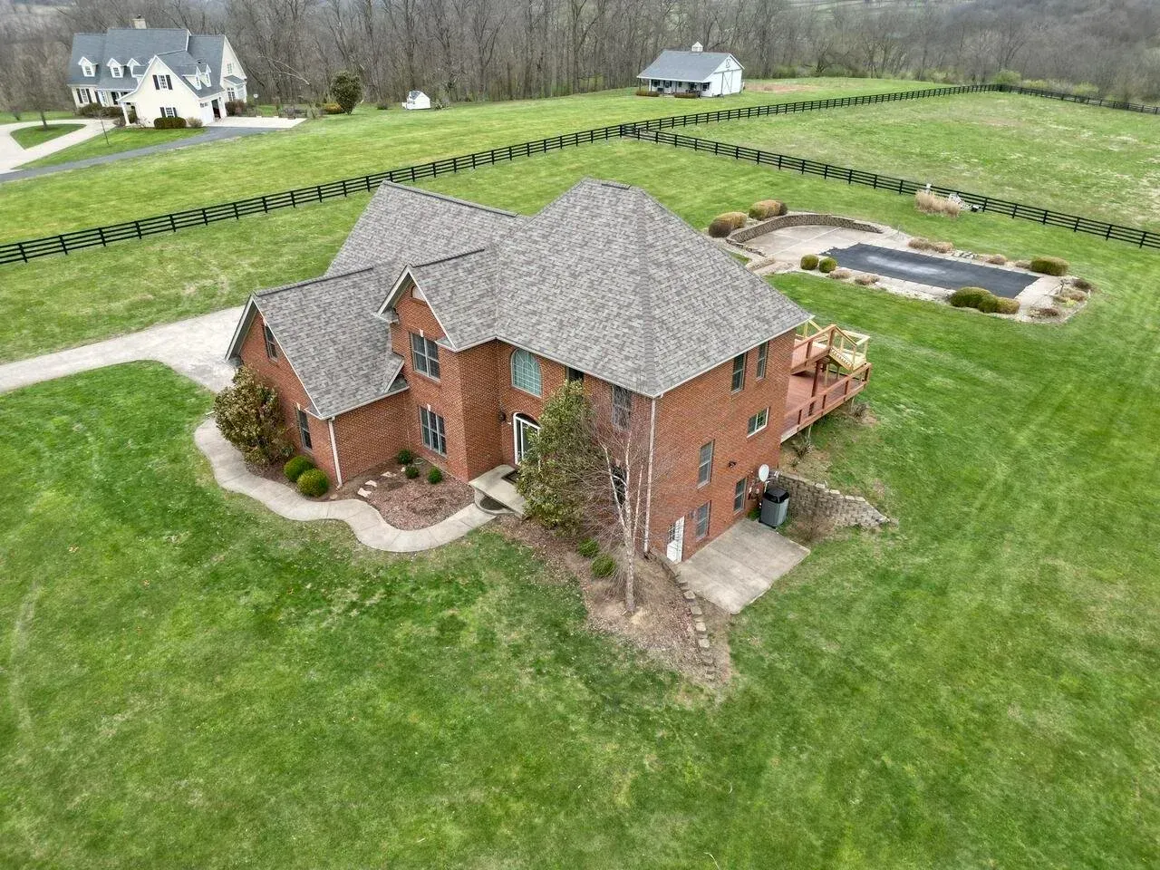 Aerial view of a red brick house with a gray roof on a large green lawn, bordered by a fence and pond.