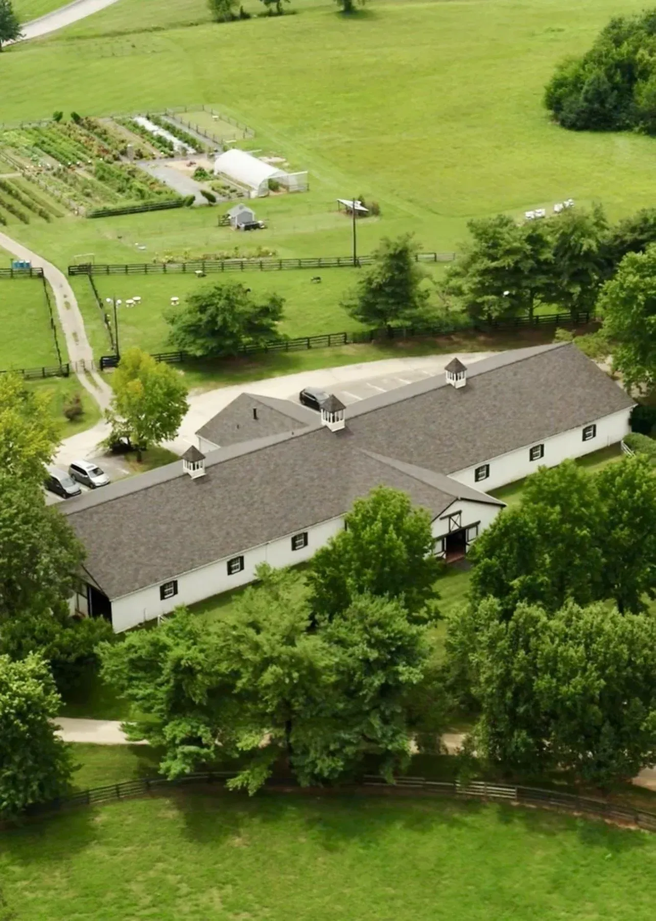 Aerial view of a long white farmhouse with a gray roof surrounded by green fields and trees.