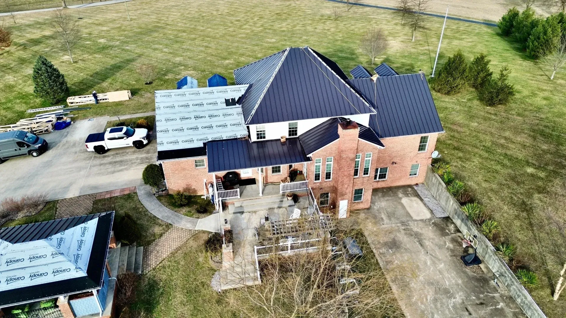 Aerial view of a large brick house with a dark roof, blue tarps, and a driveway in a grassy rural yard
