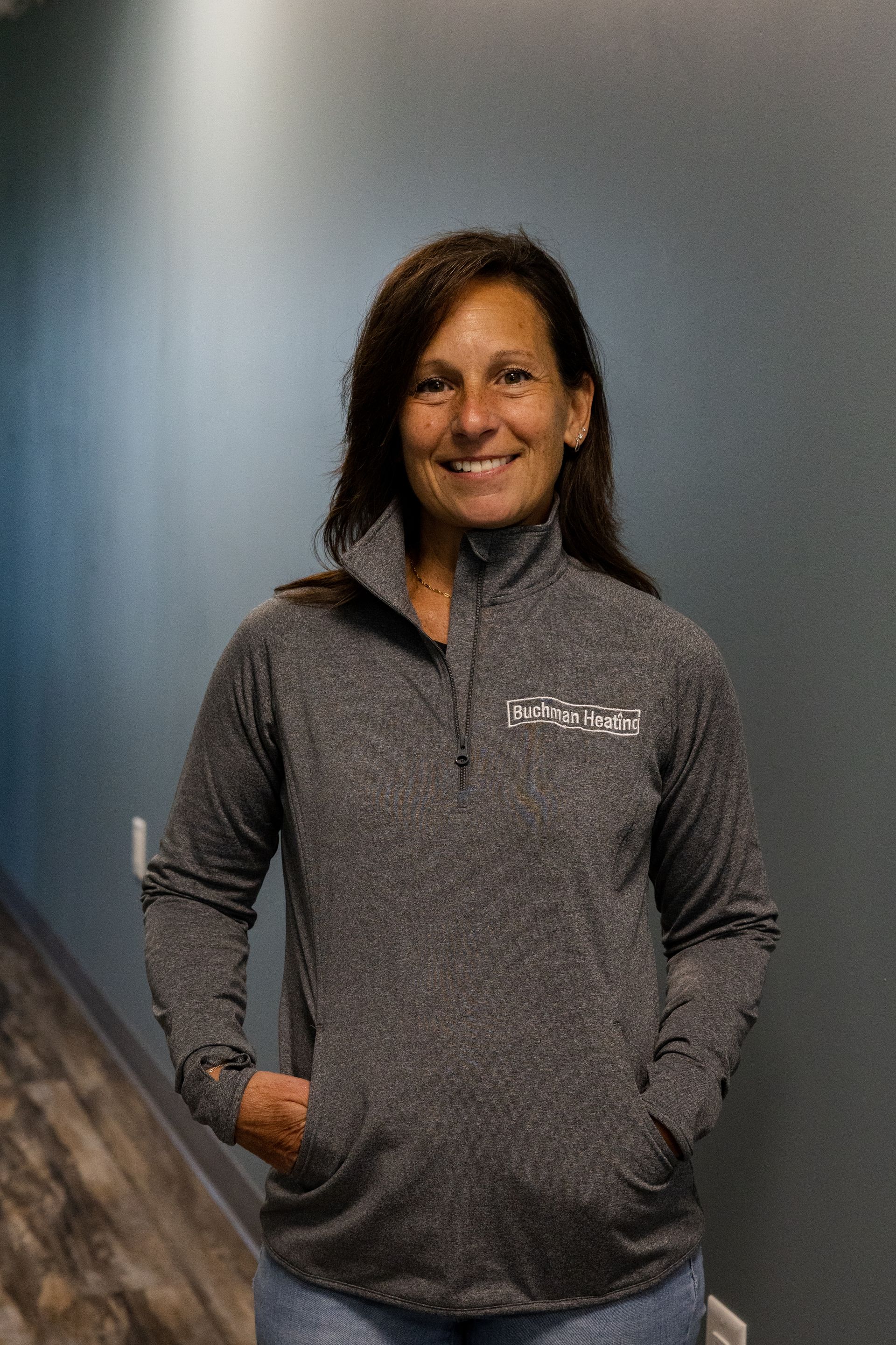 A woman wearing a gray jacket is standing in a hallway with her hands in her pockets.