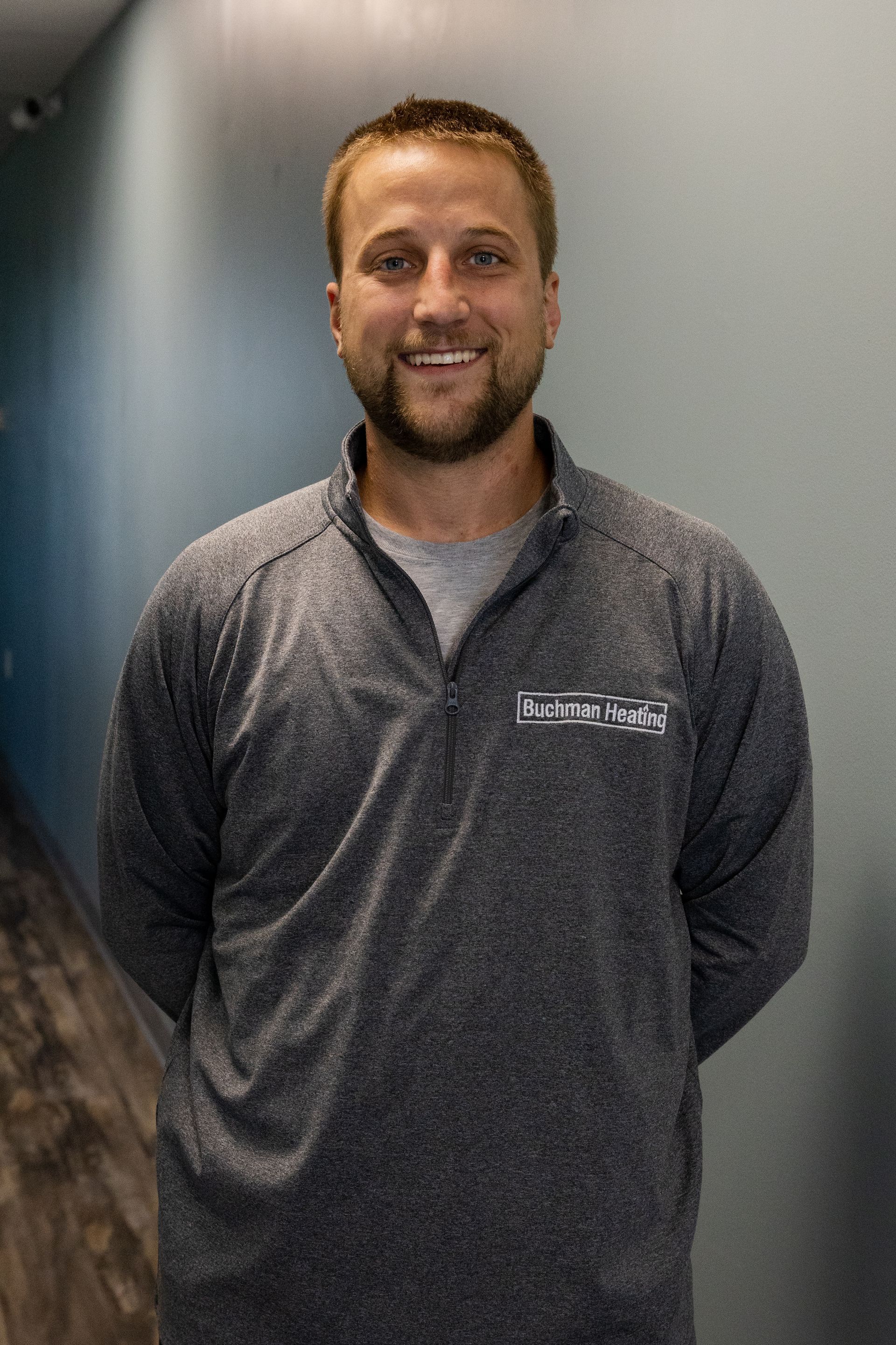 A man with a beard is standing in a hallway wearing a gray shirt.