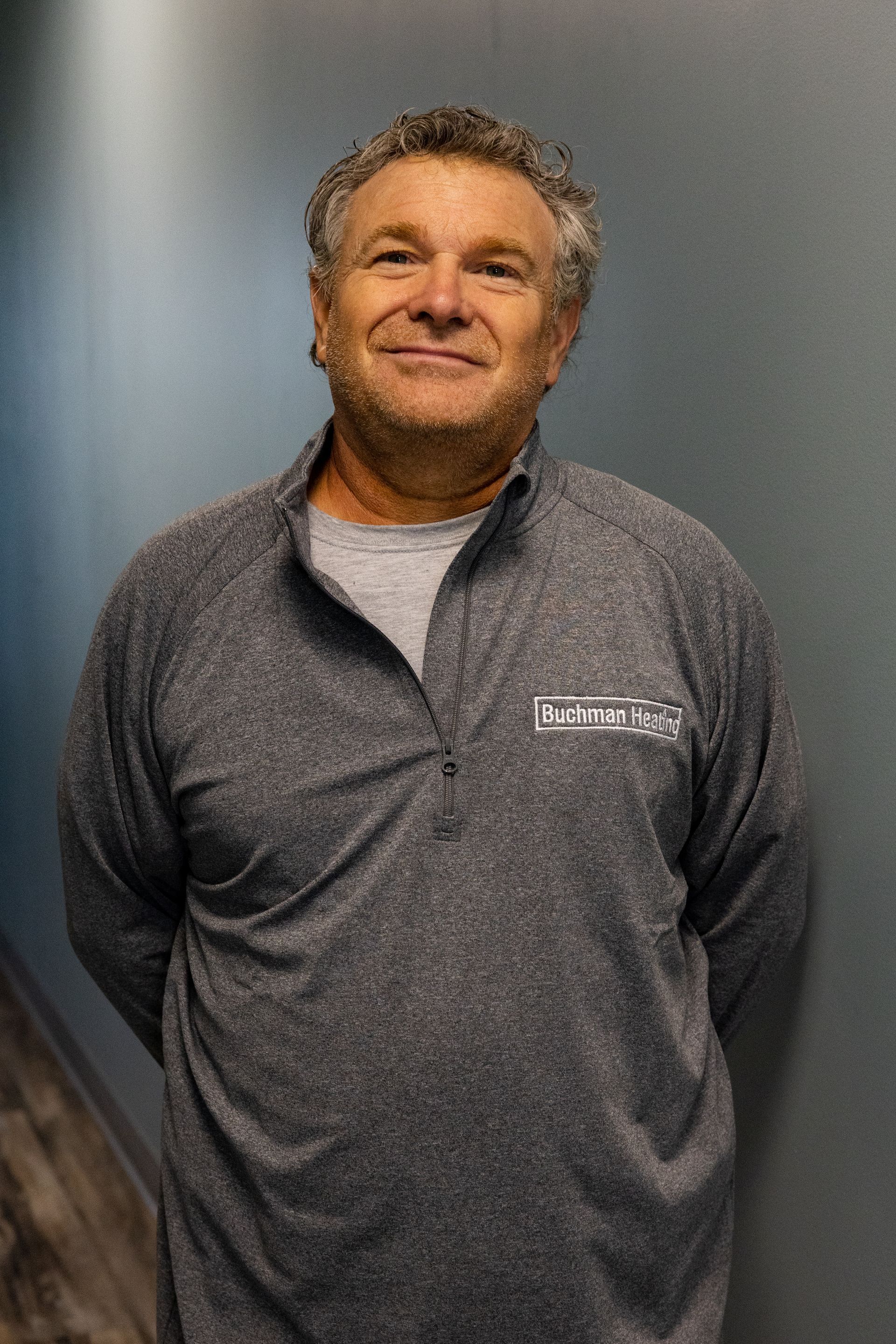 A man in a gray shirt is standing in front of a blue wall.