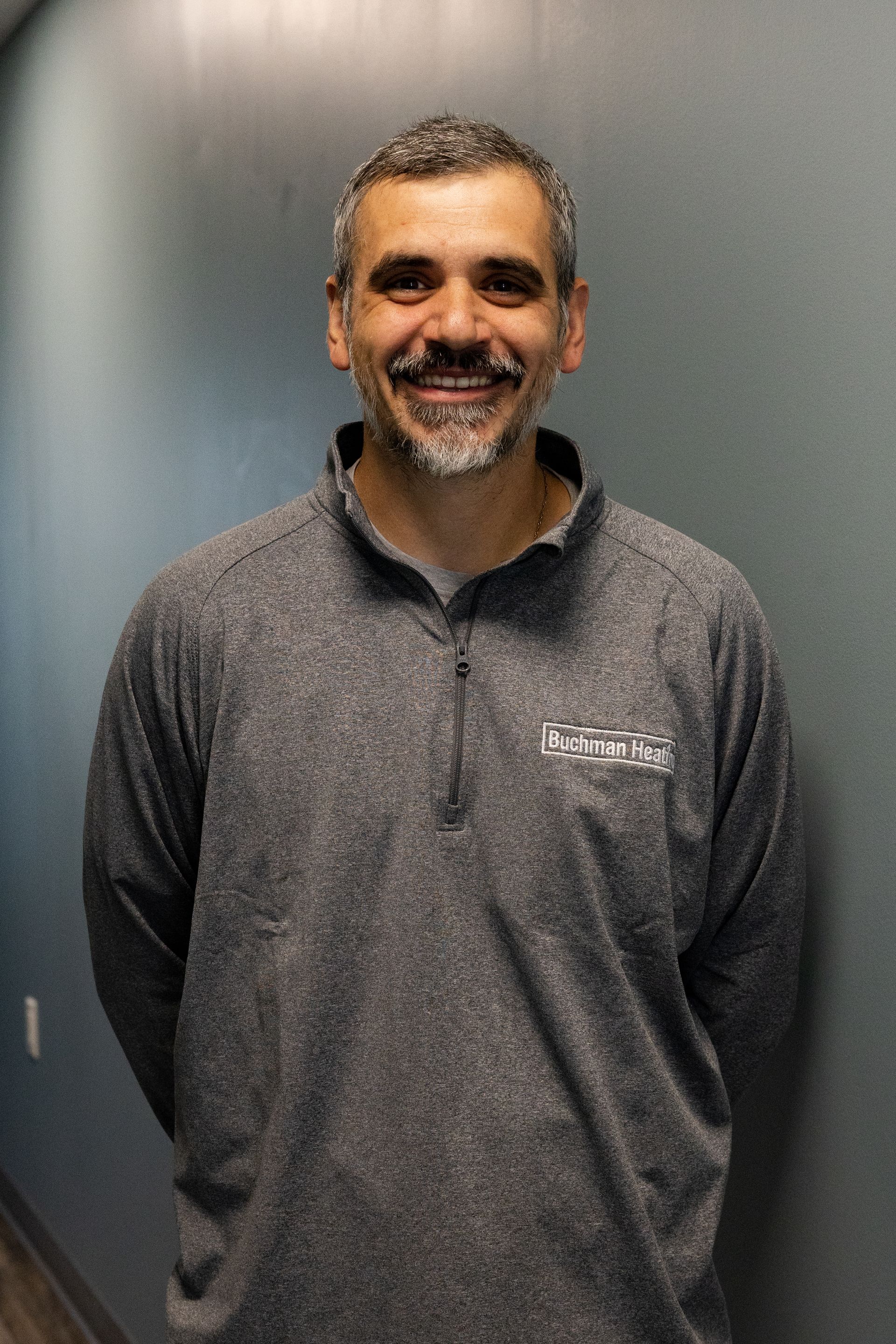 A man with a beard is wearing a gray shirt and smiling.