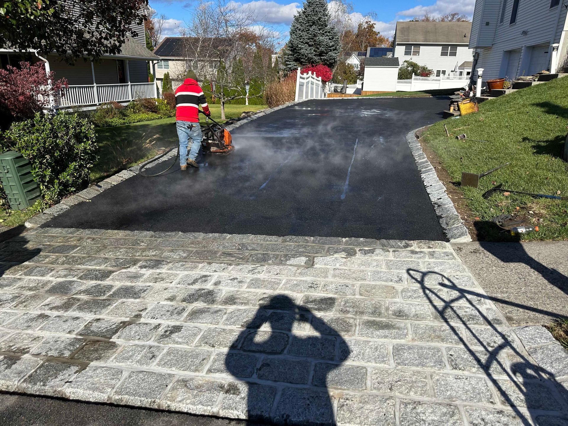 Man paving driveway with asphalt, steam rising. Cobblestone border and sidewalk. Sunny day.