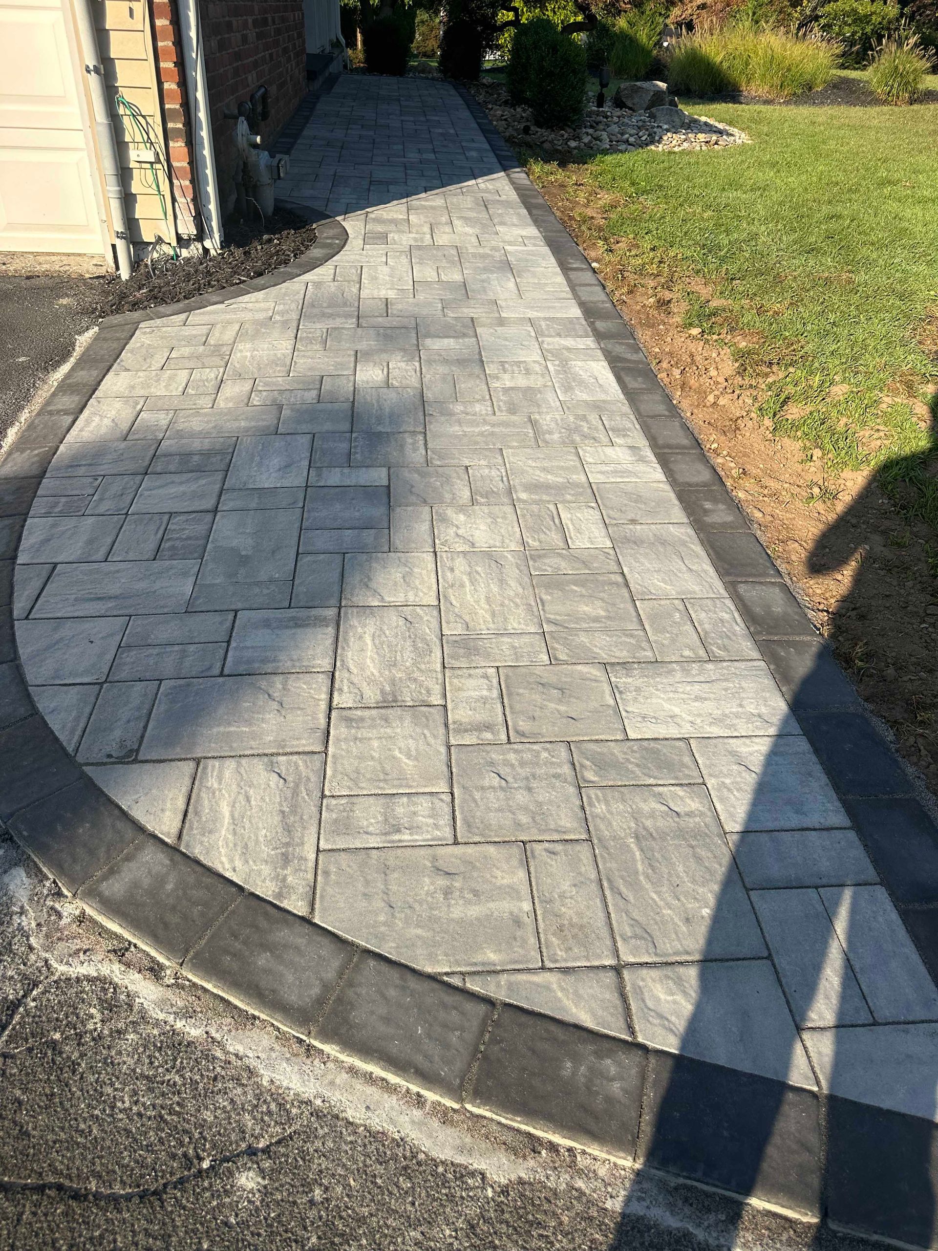 Stone paver walkway with dark border, leading to a home, adjacent to a grass lawn.